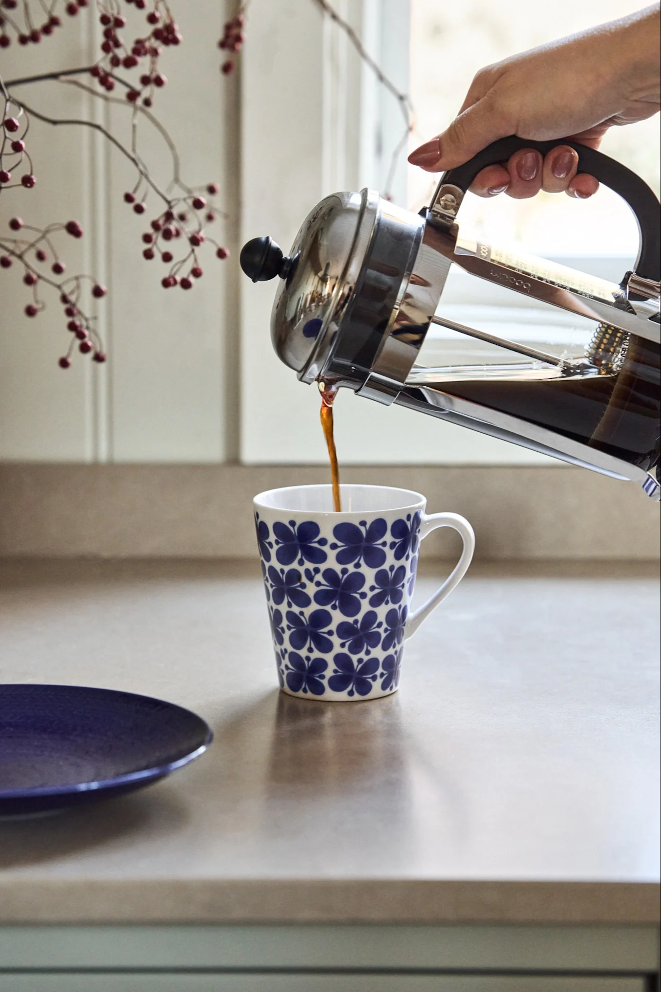Een hand schenkt koffie uit een cafetière in een witte mok met een blauw bloemmotief op een aanrecht.