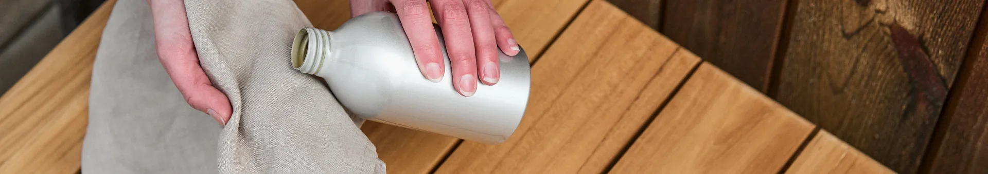 Een paar handen houden een doek en een fles teakolie vast voor het oliën van een teakhouten bank uit 1898 bij Nordic Nest. 