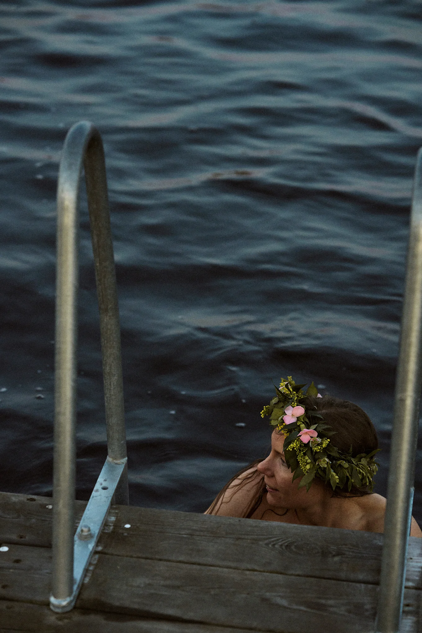 Vrouw in donker water met bloemenkrans, naast een houten steiger met metalen leuningen.