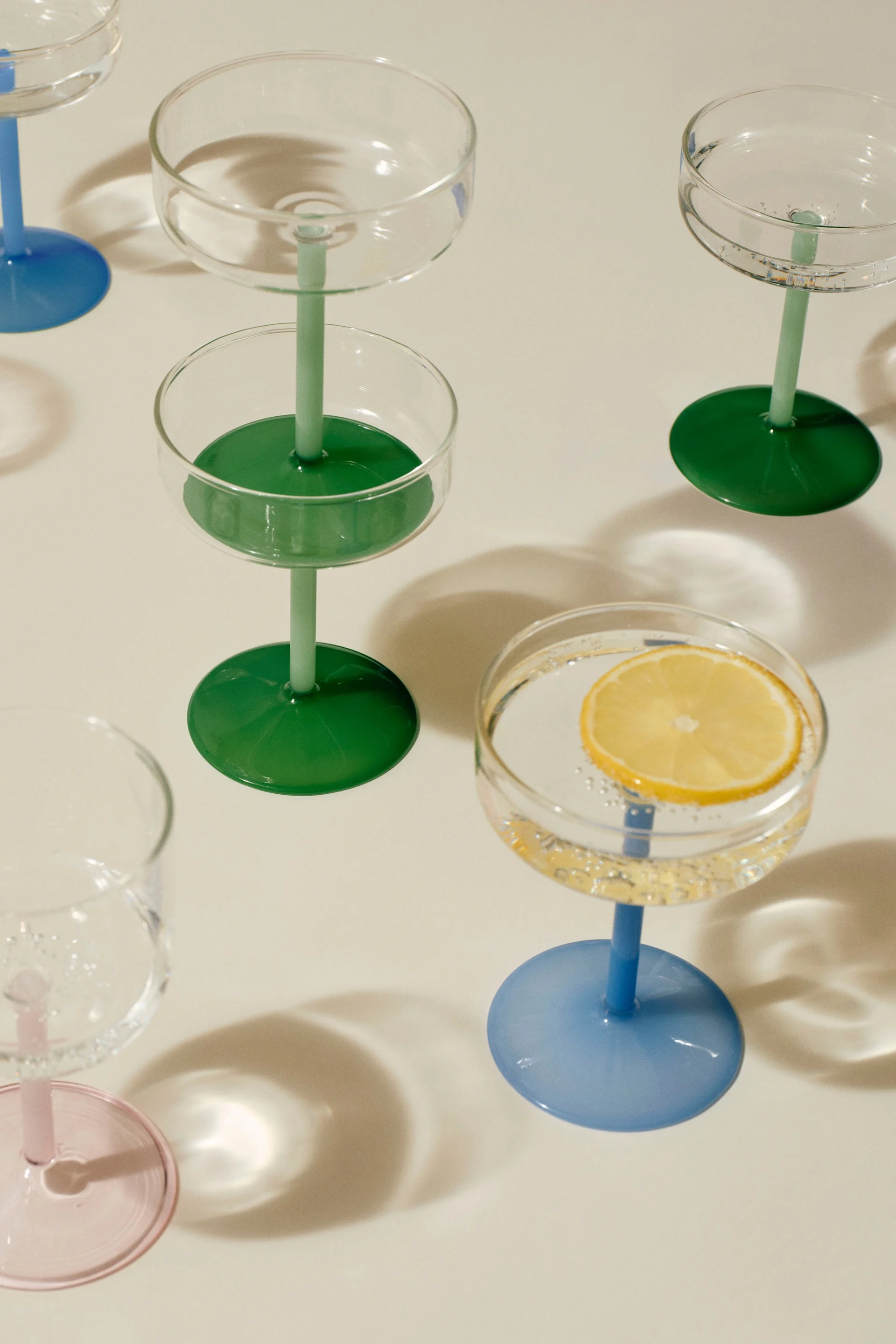 Assortiment coupeglazen met kleurrijke stelen en voetstukken. Eén glas bevat een schijfje citroen in bruisend water.
