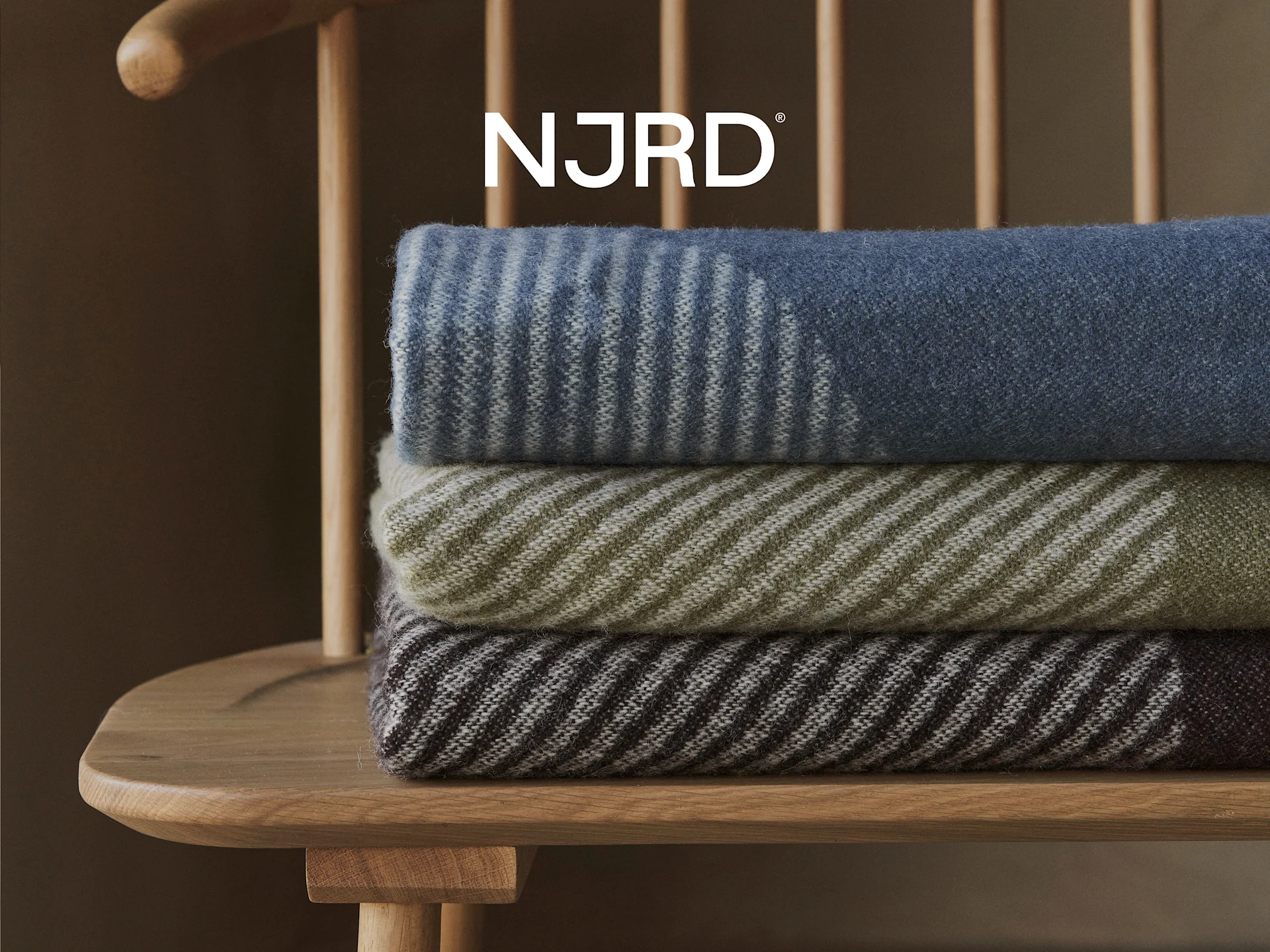 Drie gestreepte dekens in blauw, olijfgroen en bruin, gestapeld op een houten stoel met het NJRD-logo erboven.