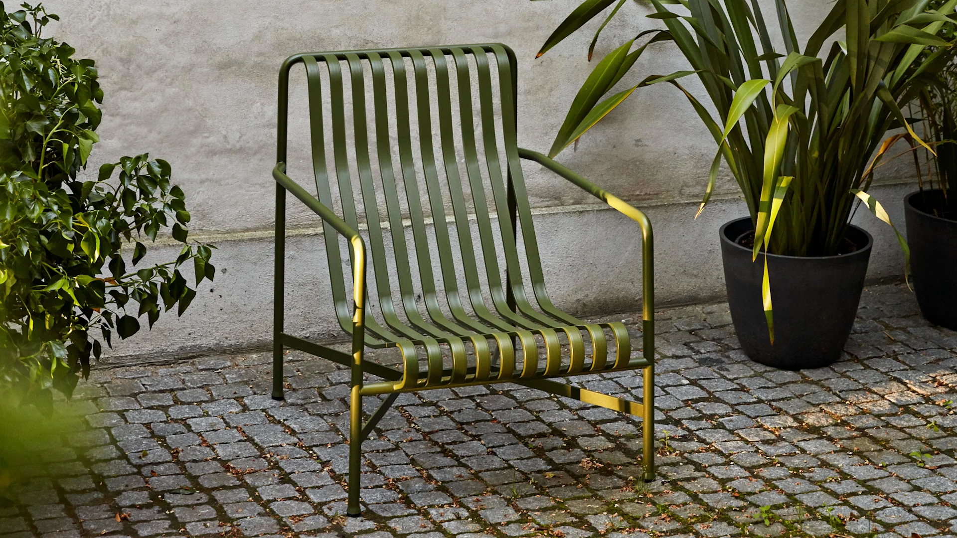 Donkergroene lamellen metalen buiten lounge stoel op een geplaveide patio, omlijst door groene planten.