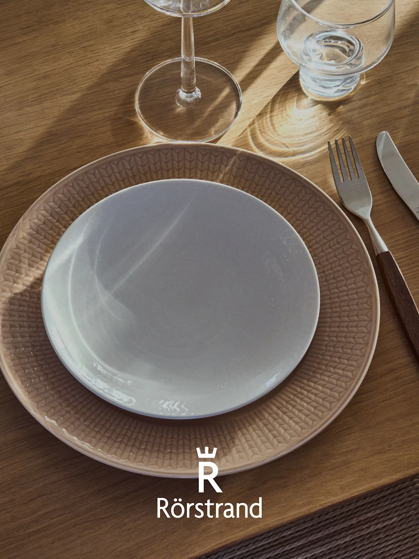 Een tafelschikking met een wit bord op een beige bord met patroon, een vork, een mes en twee glazen op een houten tafel. Rörstrand-logo.