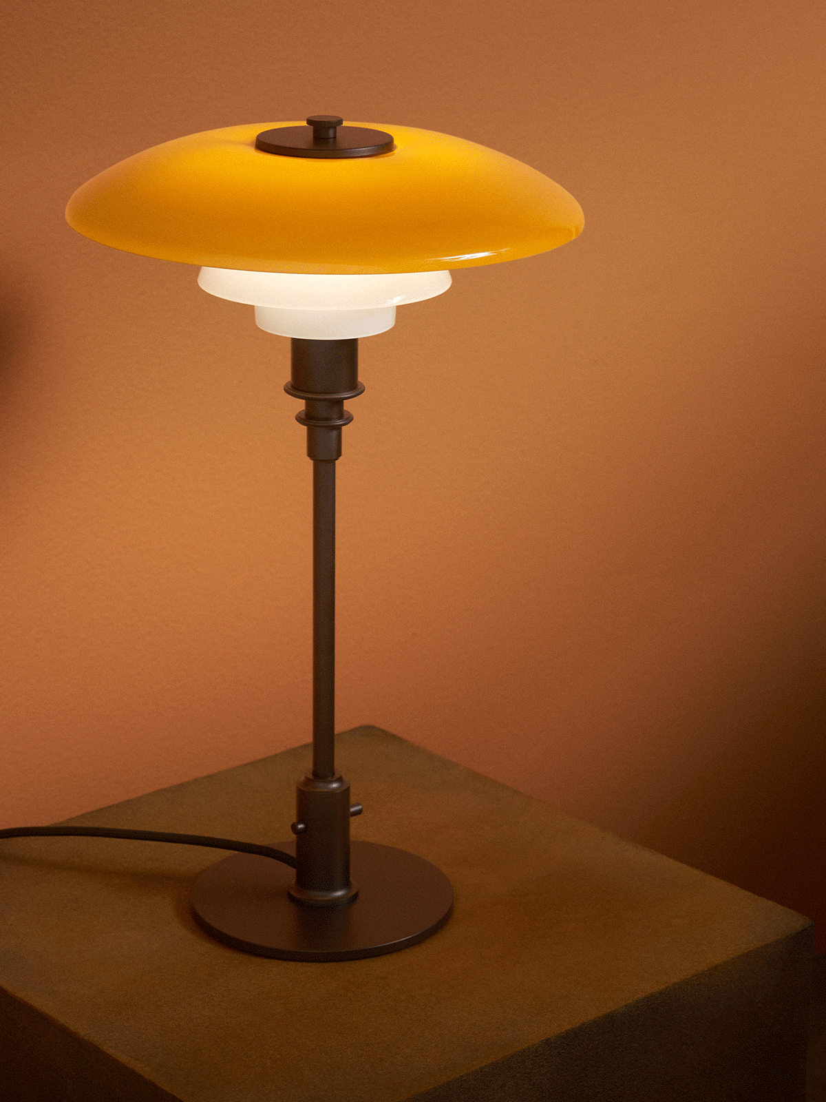 Moderne tafellamp met gele bovenkap en twee witte kappen, op een gestructureerde houten sokkel tegen een bruine muur.