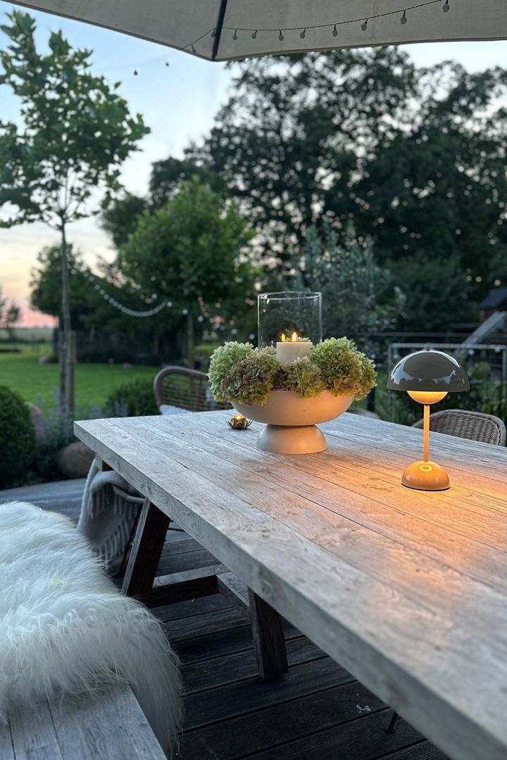 Een gezellig terras met een grote eettafel en banken, versierd met kaarsen en een Flowerpot tafellamp van &Tradition.