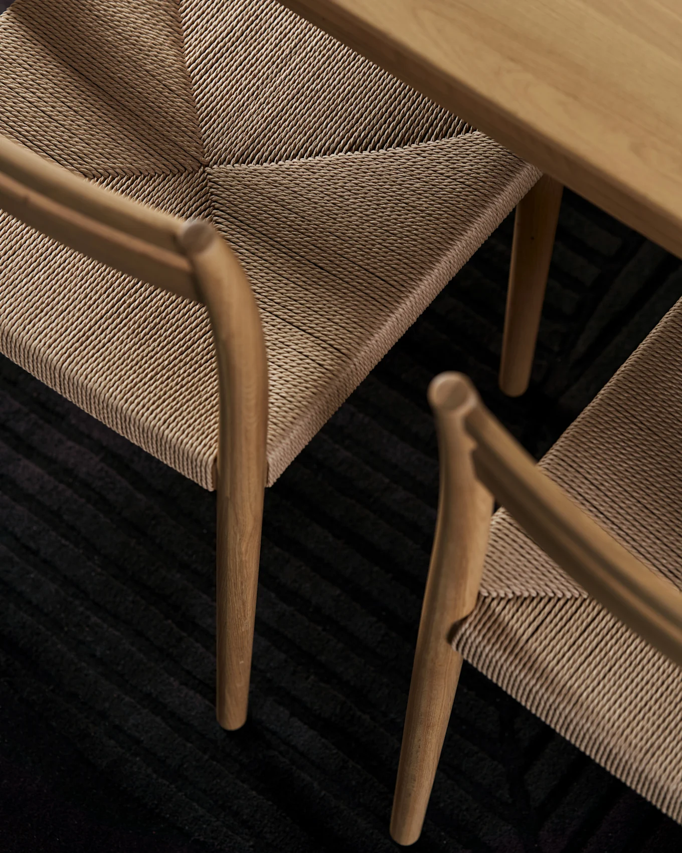 Close-up van houten stoelen met geweven touwzittingen onder een houten tafel op een donker geribbeld tapijt.