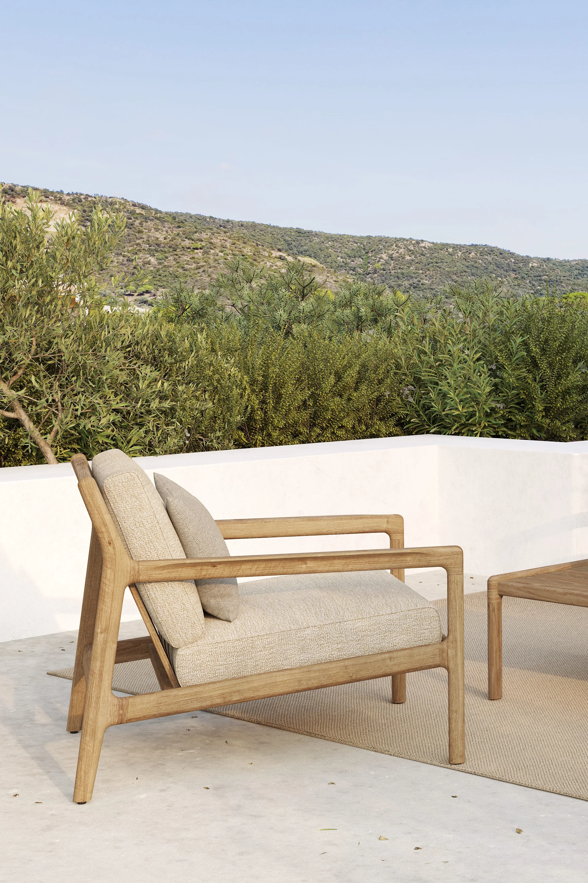 Houten buitenfauteuil met lichte kussens op een patio, met groene heuvels en een blauwe lucht op de achtergrond.