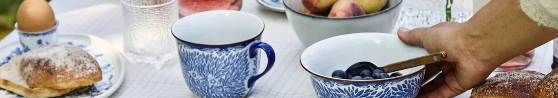 Een ontbijttafel met ei, broodjes, bosbessen en perziken. Een hand pakt een kom met bosbessen.