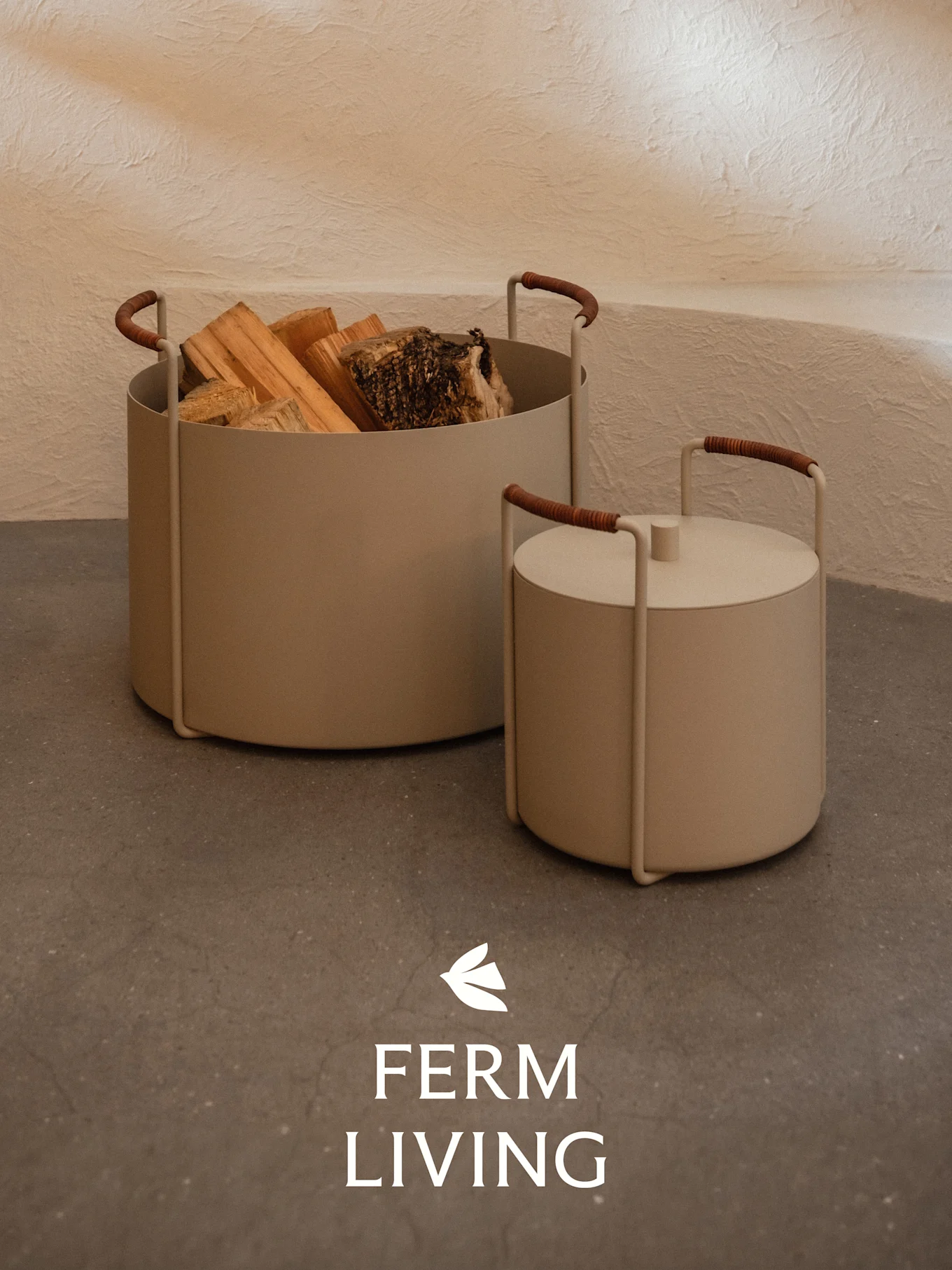 Twee beige cilindrische containers, één met brandhout en de andere met deksel, met bruin omwikkelde handvatten op een betonnen vloer.