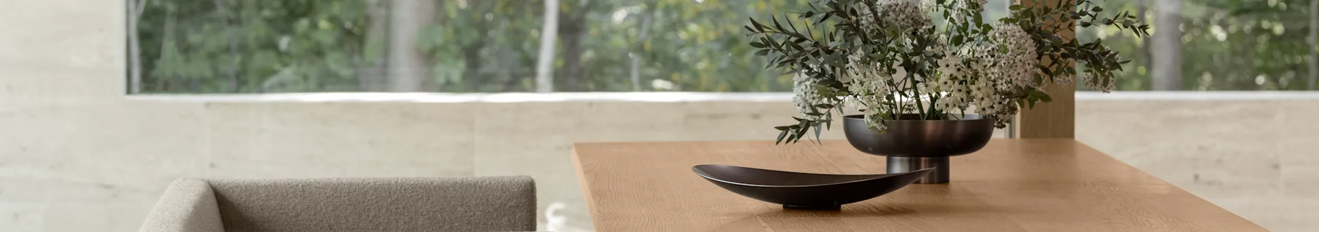 Moderne houten eettafel met een decoratieve donkere kom en een vaas met witte en groene bloemen, met een wazig groen uitzicht naar buiten door een groot raam.