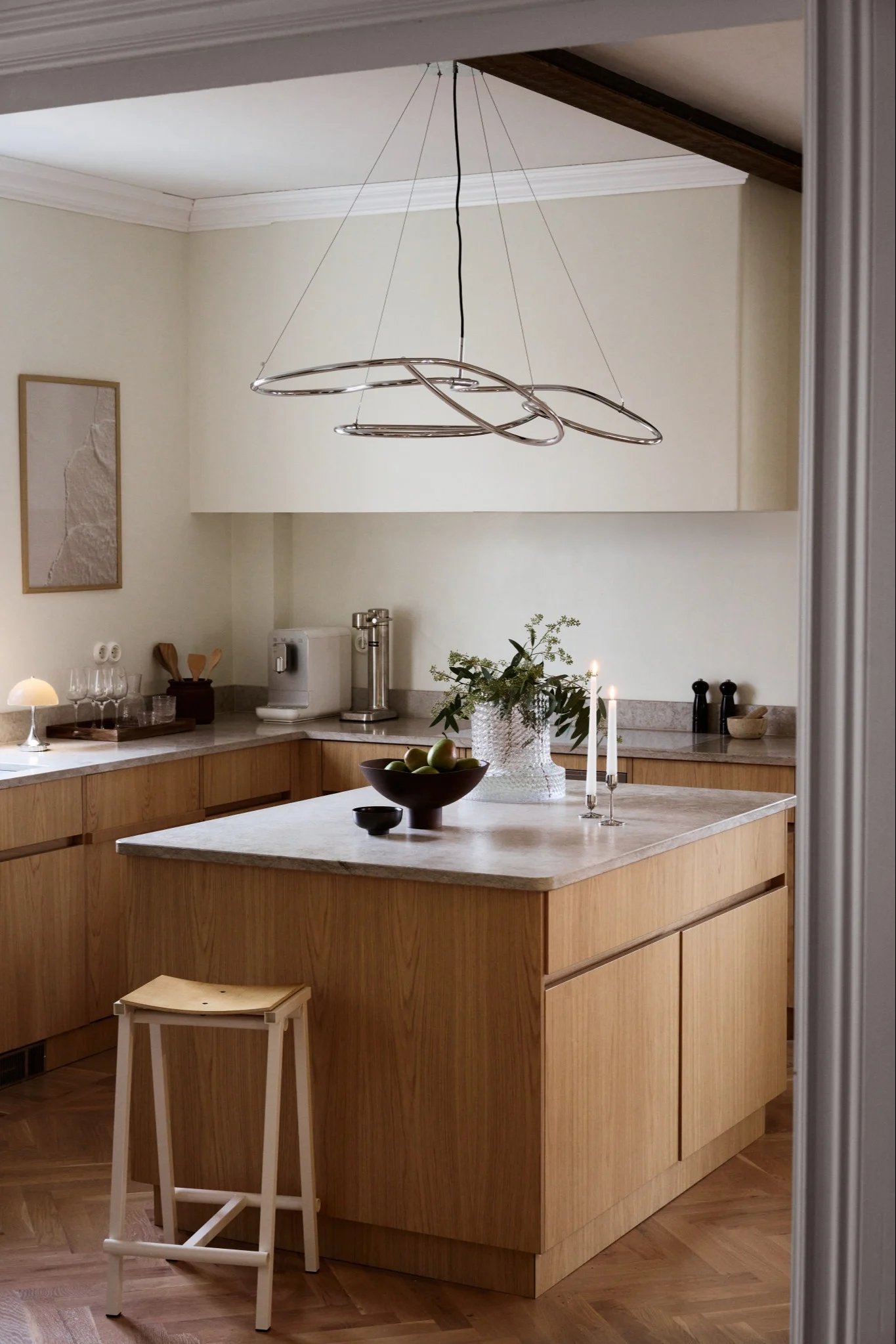 Moderne keuken met lichte houten kasten, marmeren werkbladen, eiland en sculpturale chromen kroonluchter.