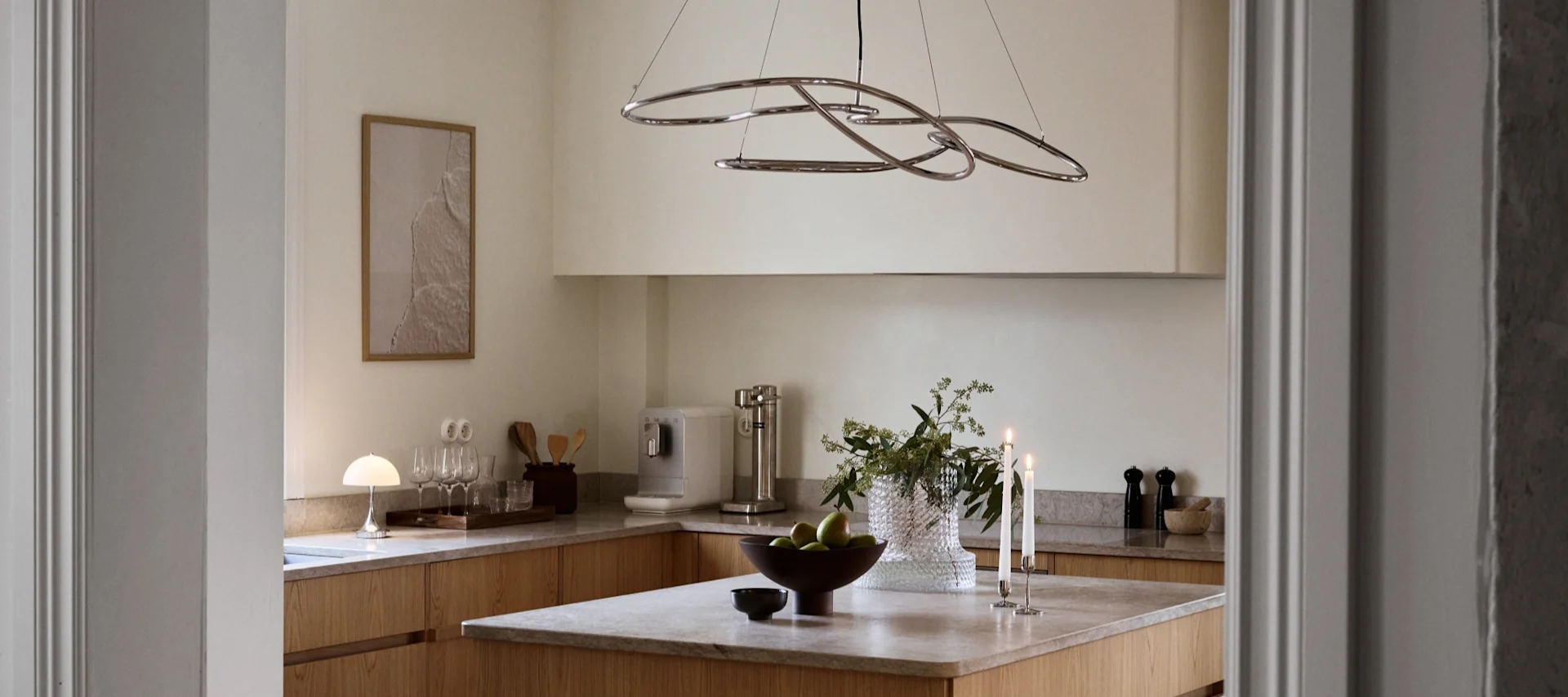 Moderne keuken met lichte houten kasten, eiland met groene appels, brandende kaarsen en sculpturale chromen kroonluchter.