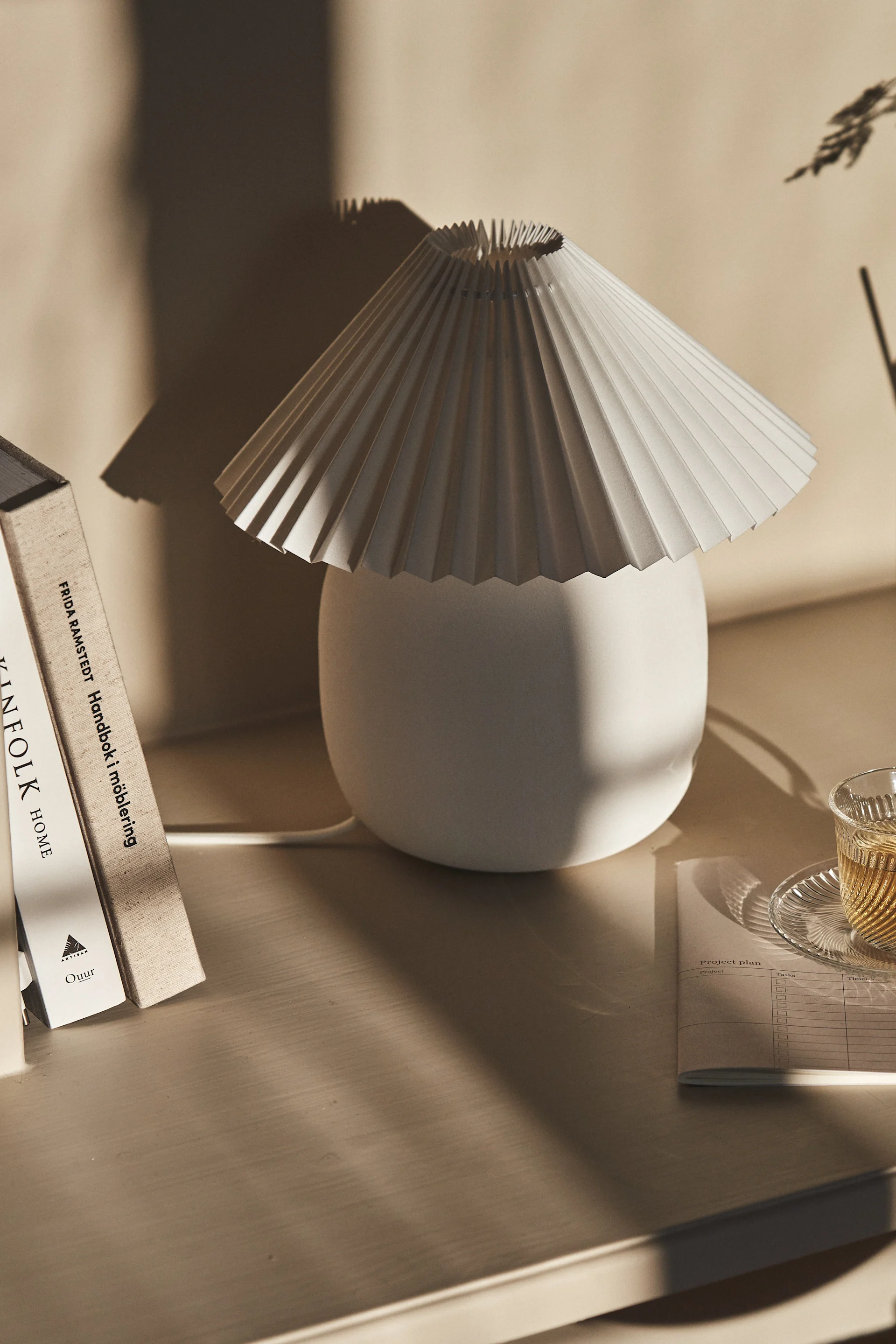 Een geplooide witte tafellamp naast boeken en een kop thee op een licht houten bureau, met zachte schaduwen.