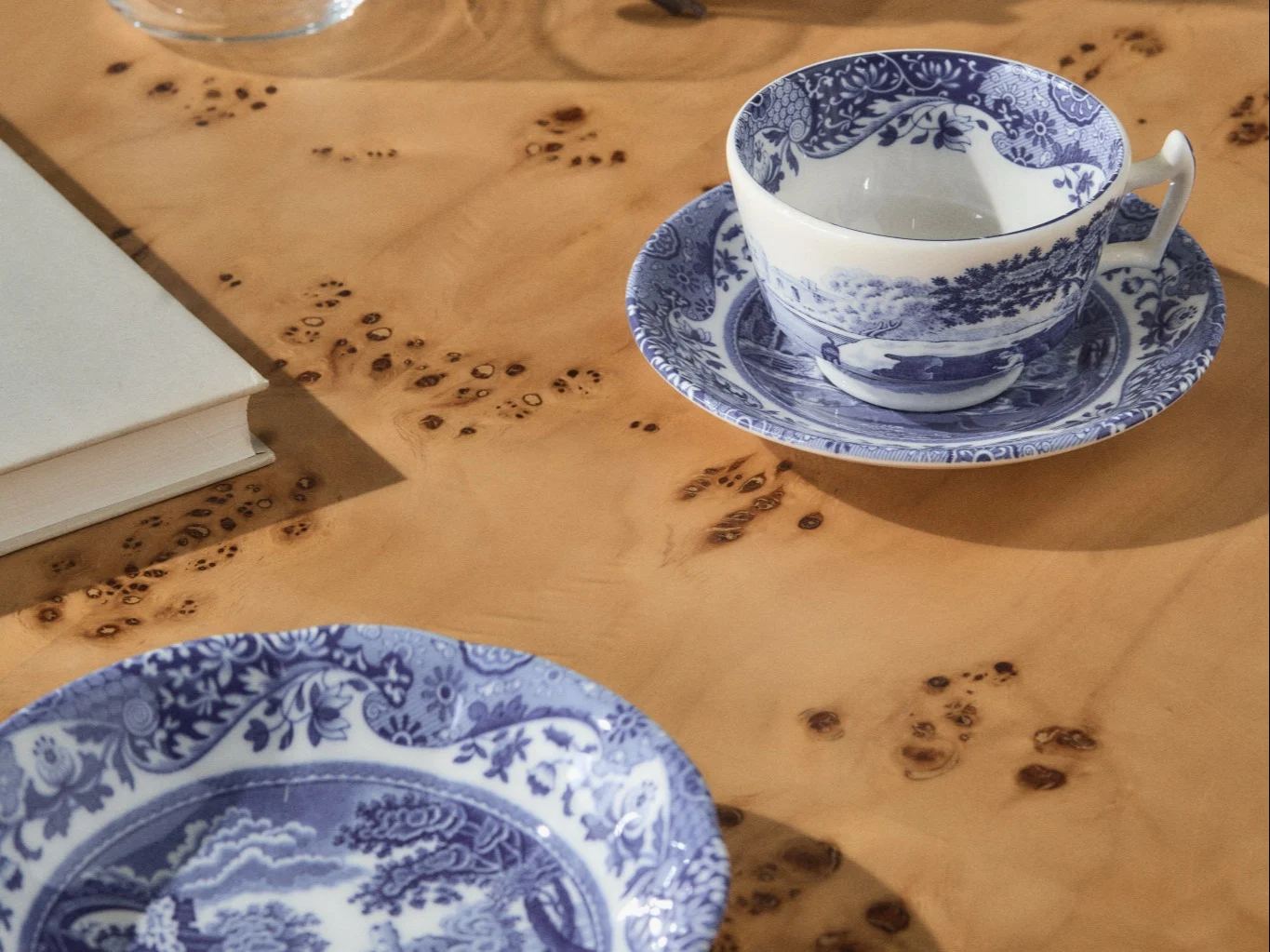 Blauw-wit gedessineerd theekopje en schoteltje met een bijpassend bord op een lichtbruine wortelhouten tafel, een wit boek zichtbaar op de achtergrond.