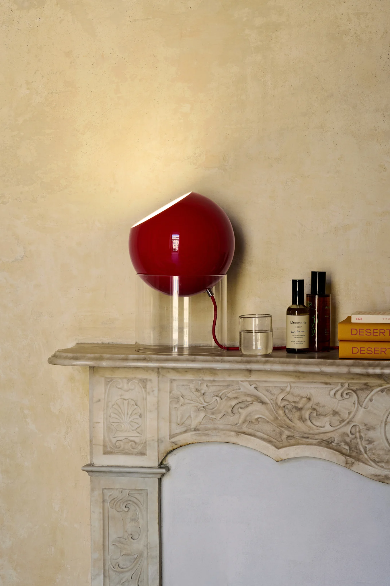 Rode bolvormige lamp met doorschijnende voet op marmeren schoorsteenmantel, naast glas water, twee flessen en twee gele boeken.