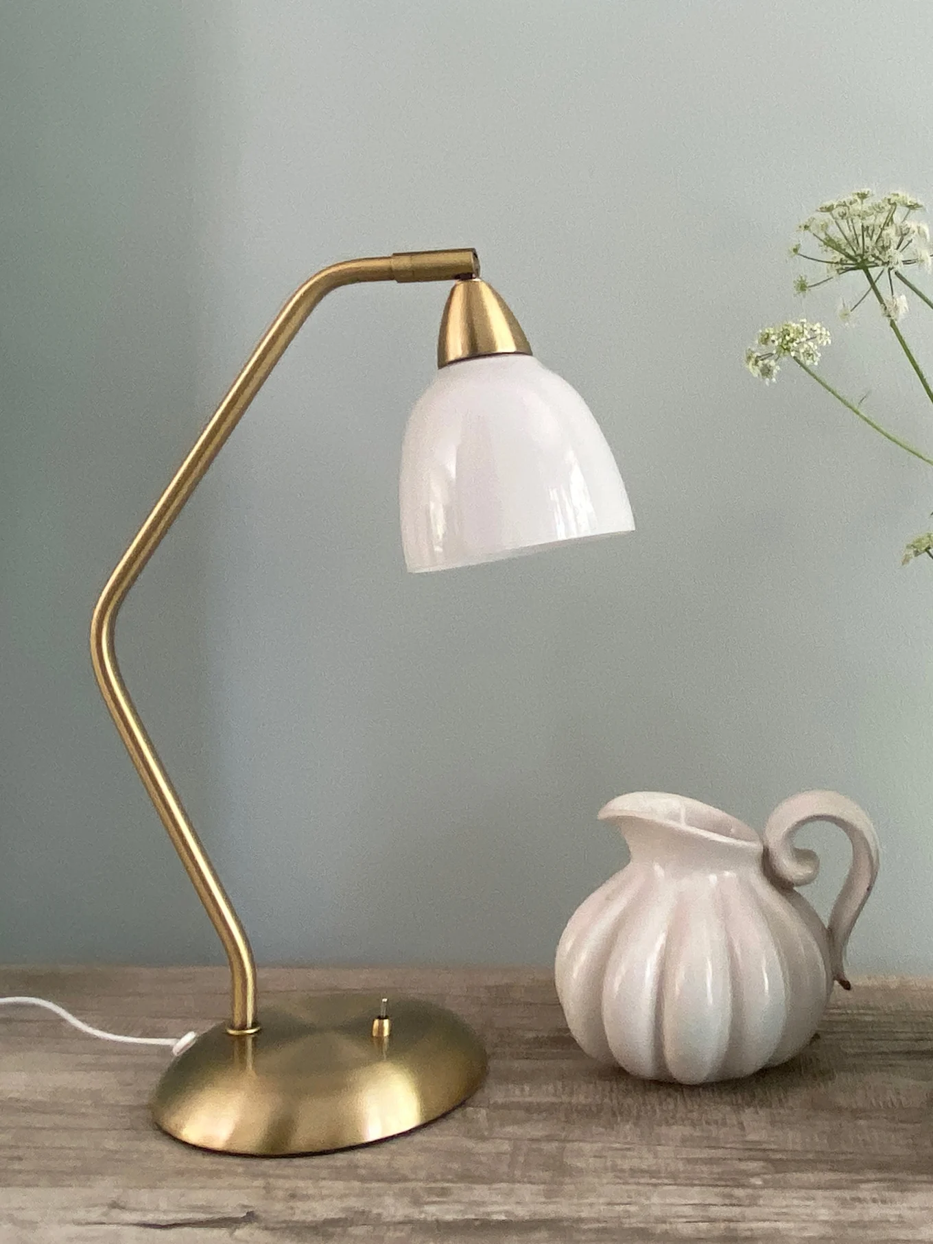 Een messing tafellamp met een witte glazen kap staat naast een witte geribbelde keramische kan op een houten oppervlak.