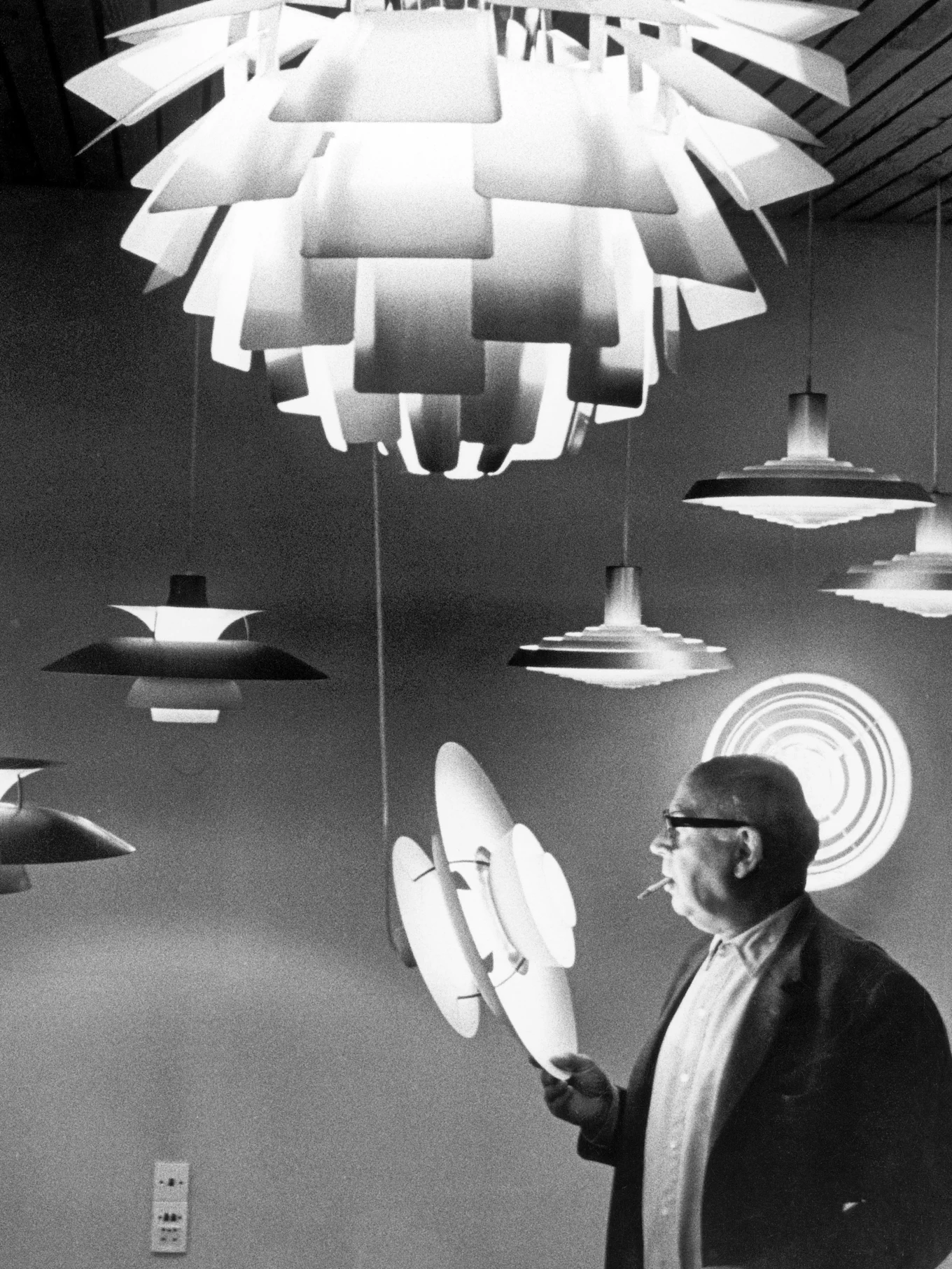 Man met bril en sigaret inspecteert meerdere gelaagde, moderne lampen in zwart-wit.