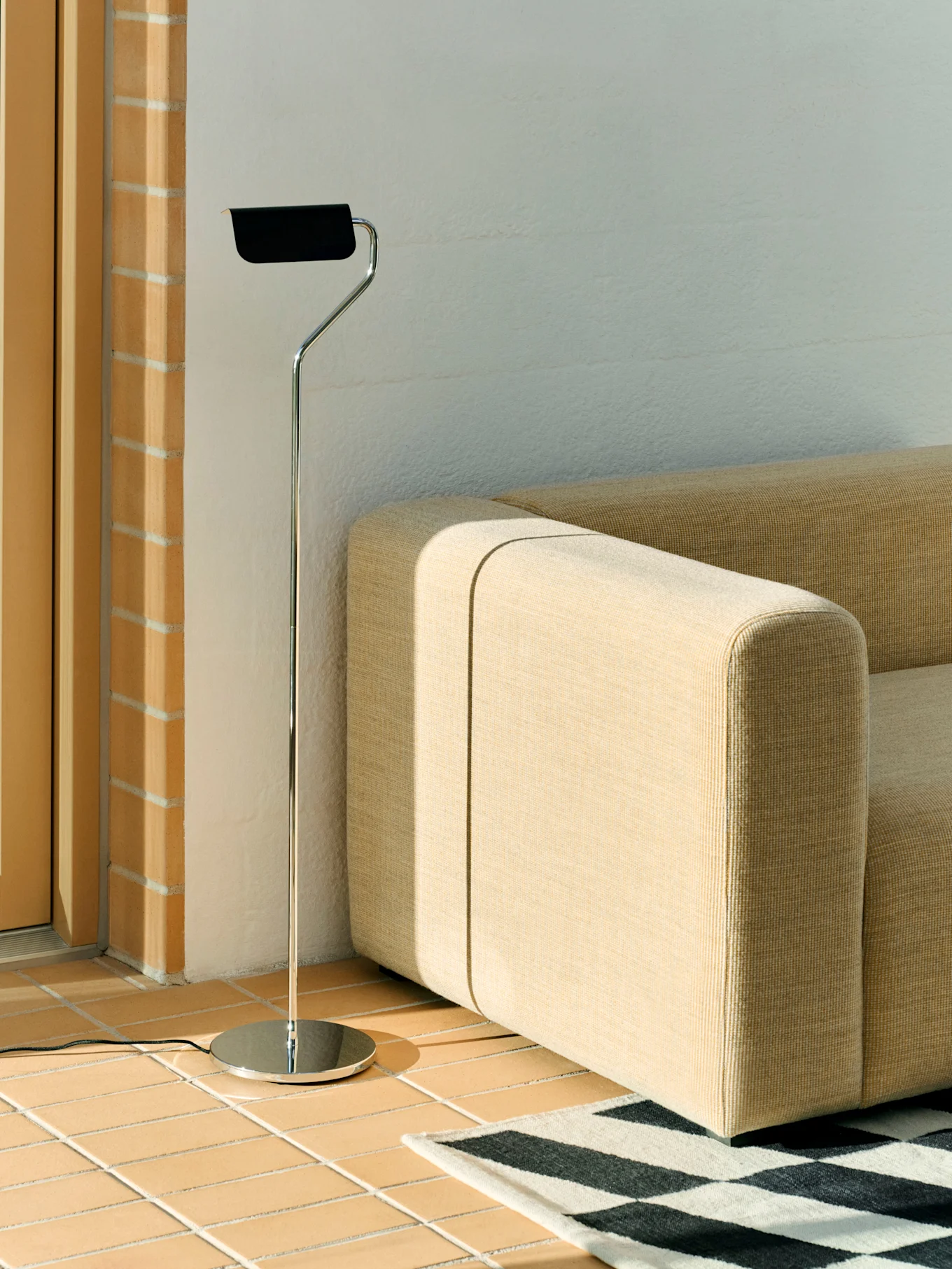 Een chromen vloerlamp met zwarte kap staat naast een beige modulaire bank en een zwart-wit geometrisch tapijt op een tegelvloer.