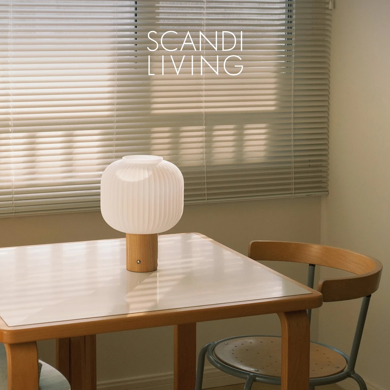 Witte geribbelde tafellamp met houten voet op een wit blad tafel en een stoel, voor raamjaloezieën. Tekst: SCANDI LIVING.