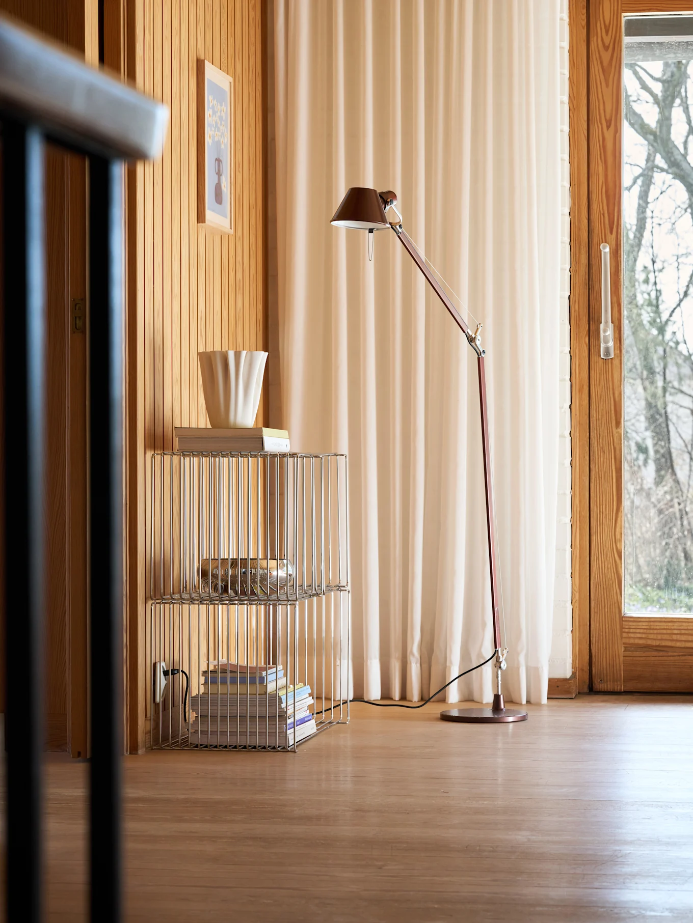 Een gezellige kamer met houten wanden en vloer, een rode vloerlamp, een chromen rek met boeken en een geribbelde witte vaas, en een raam met lichte gordijnen.