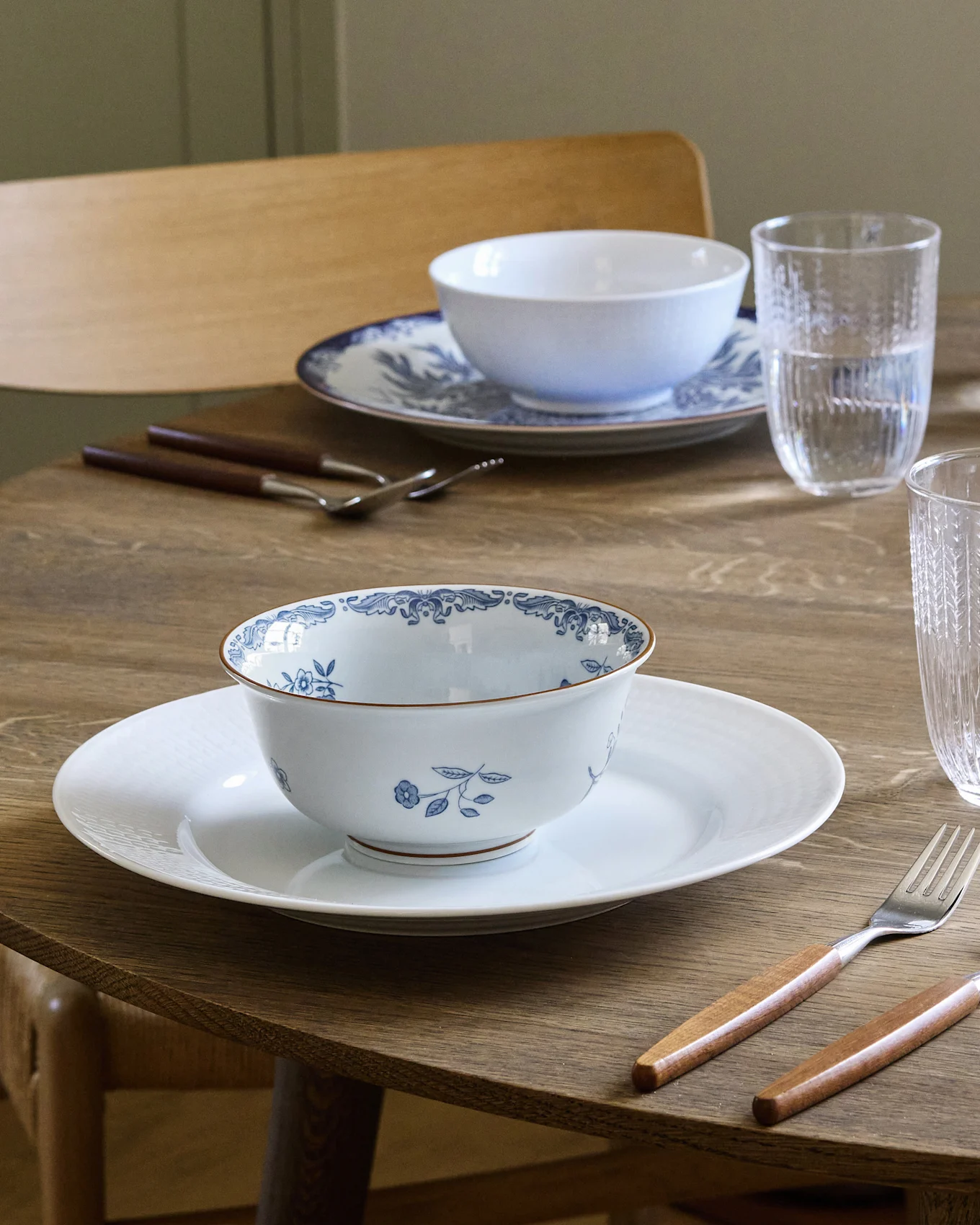 Eettafel gedekt voor twee met wit en blauw gedessineerde kommen op witte borden, bestek met houten handvatten en waterglazen.
