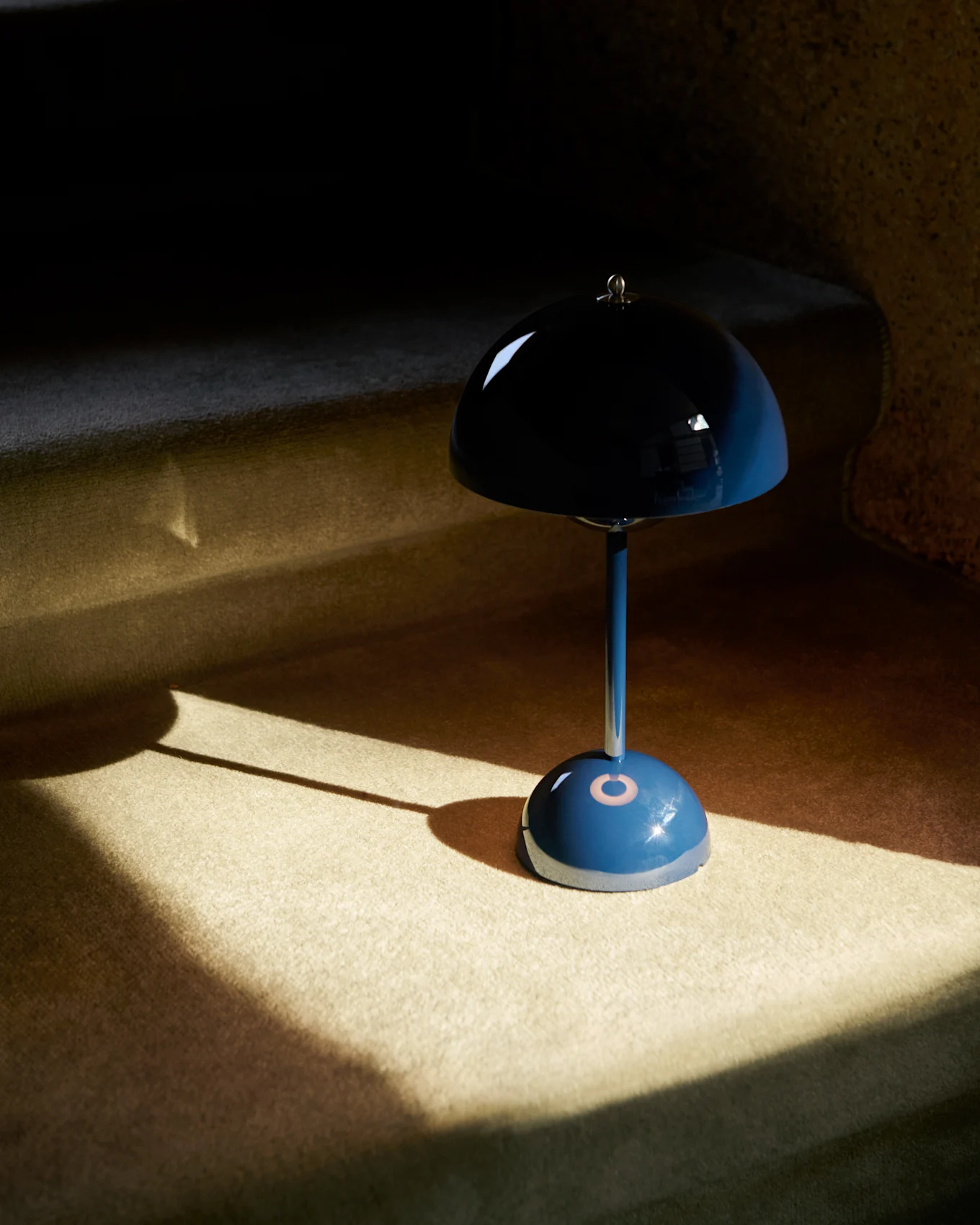 Een blauwe paddestoelvormige tafellamp met een reflecterende kap staat op een vloerbedekking, deels verlicht door zonlicht.