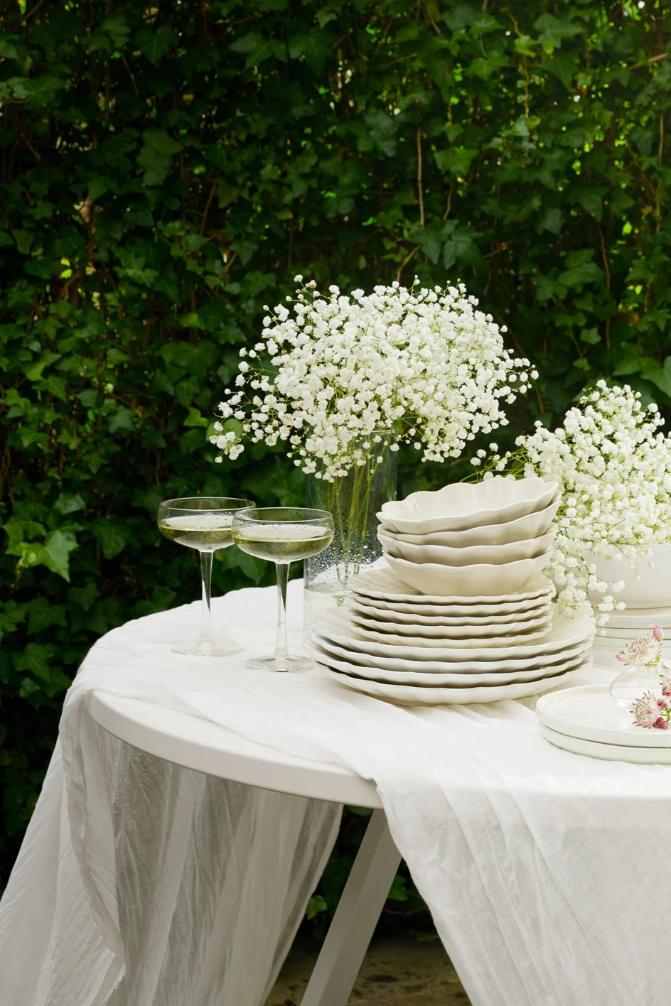 Buitentafel gedekt met wit servies, champagneglazen en gipskruidbloemen tegen groene bladeren.