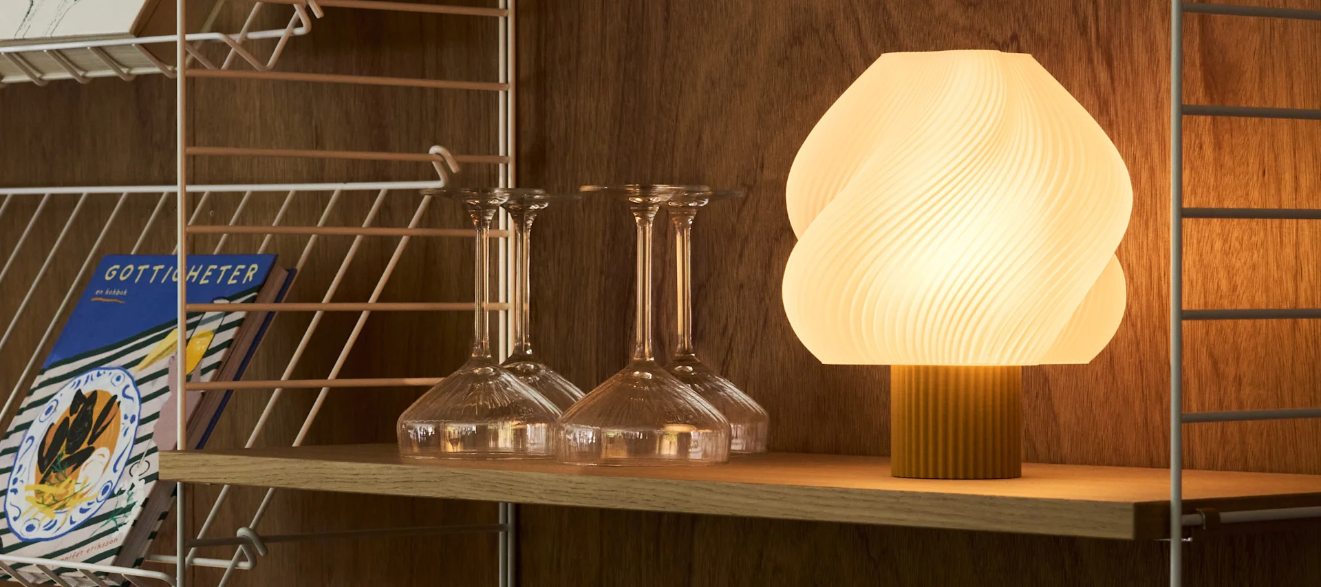Een verlichte, geribbelde ovale lamp en diverse heldere glazen staan op een houten barwagen tegen een warme, getextureerde muur.