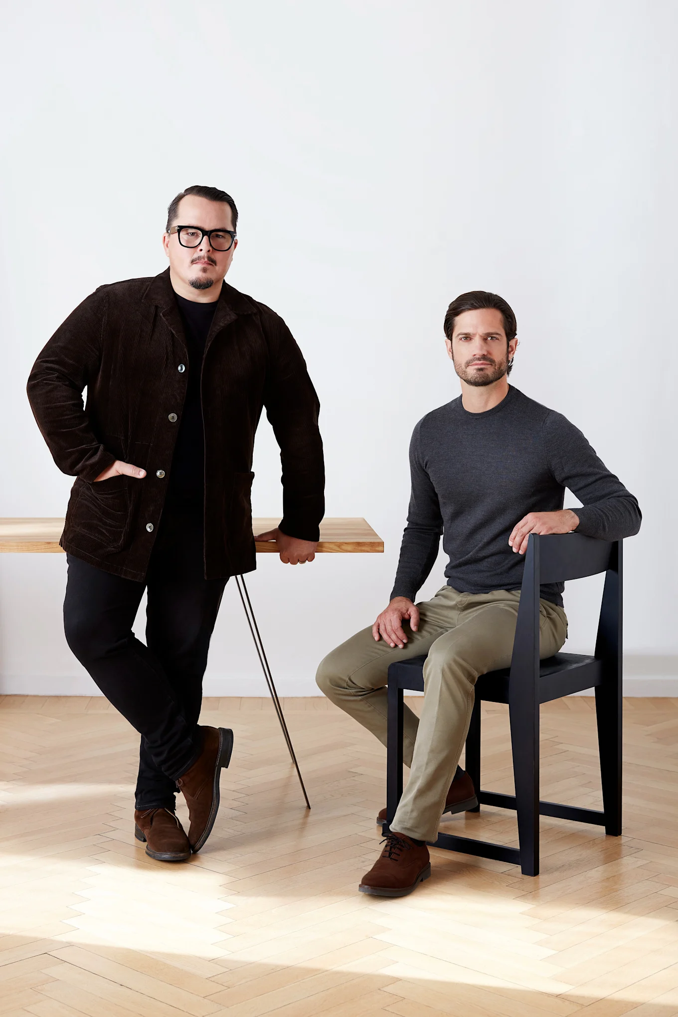 Twee mannen, één staat bij een houten tafel in een bruine corduroy jas, de ander zit op een zwarte stoel in een grijs overhemd.