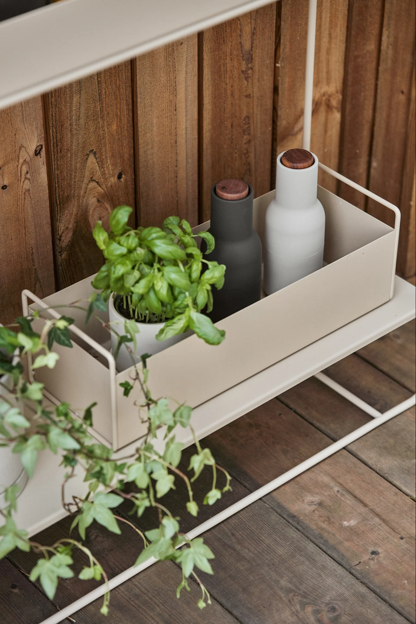 Een basilicumplant en twee minimalistische flessen (donkergrijs en lichtgrijs) met houten doppen staan op een lichtbeige metalen plank tegen een houten muur.