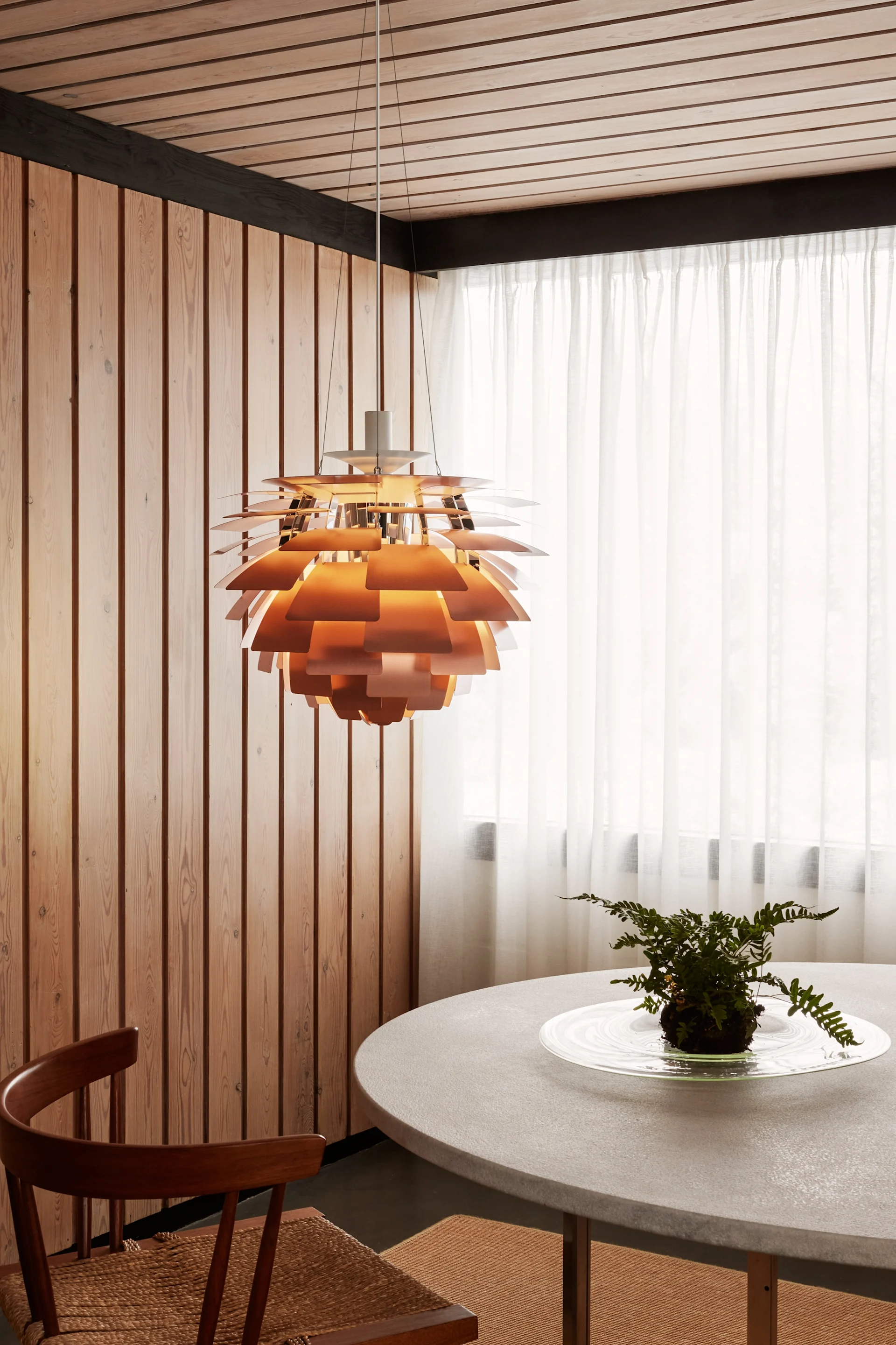 Een koperkleurige gelaagde hanglamp hangt boven een ronde grijze tafel met een varen, naast houten wanden en transparante witte gordijnen.