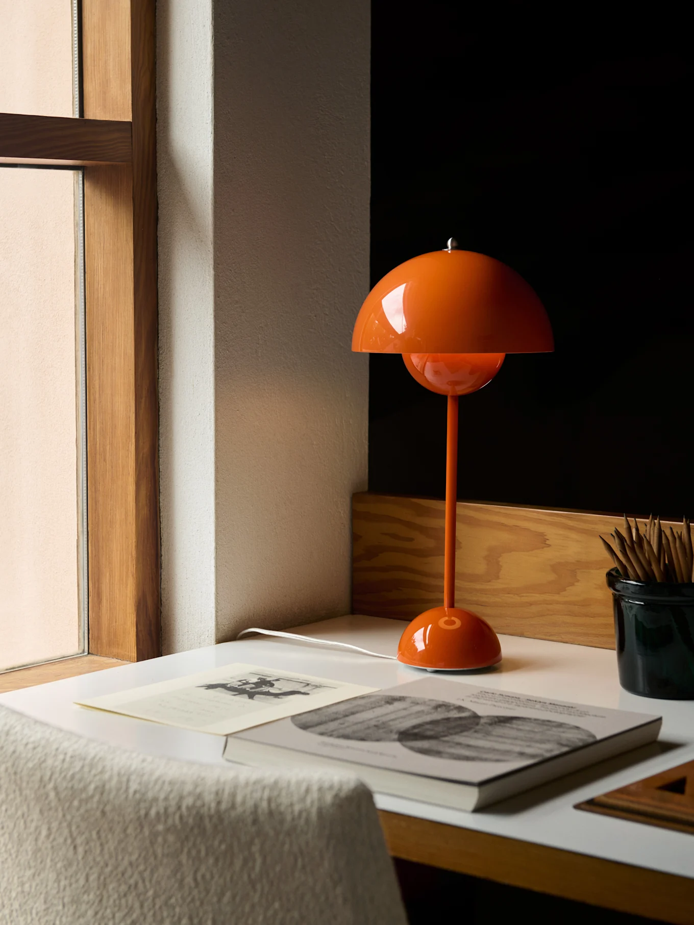 Een oranje paddestoelvormige tafellamp op een wit bureau naast een raam, met boeken en potloden.