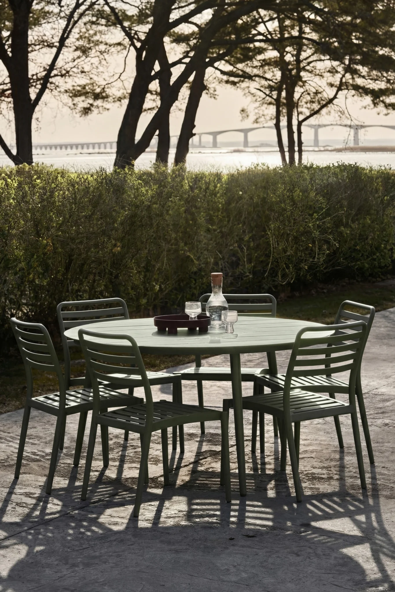 Groene tuineetset met tafel, vijf stoelen, waterfles en glazen op terras. Verre brug over water.