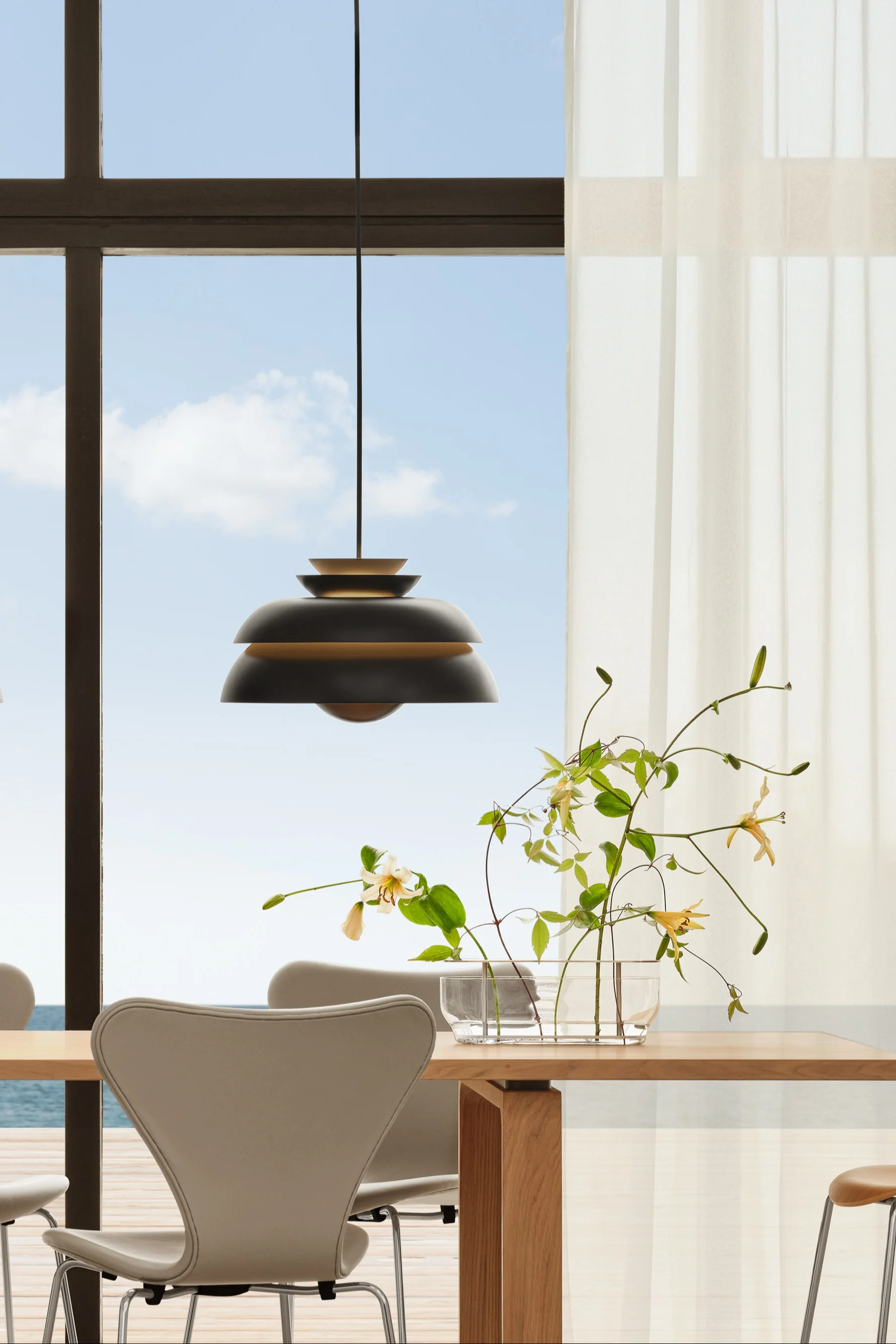 Eetkamer: houten tafel, stoelen, donkere hanglampen. Bloemen. Raam, vitrages, uitzicht op zee.