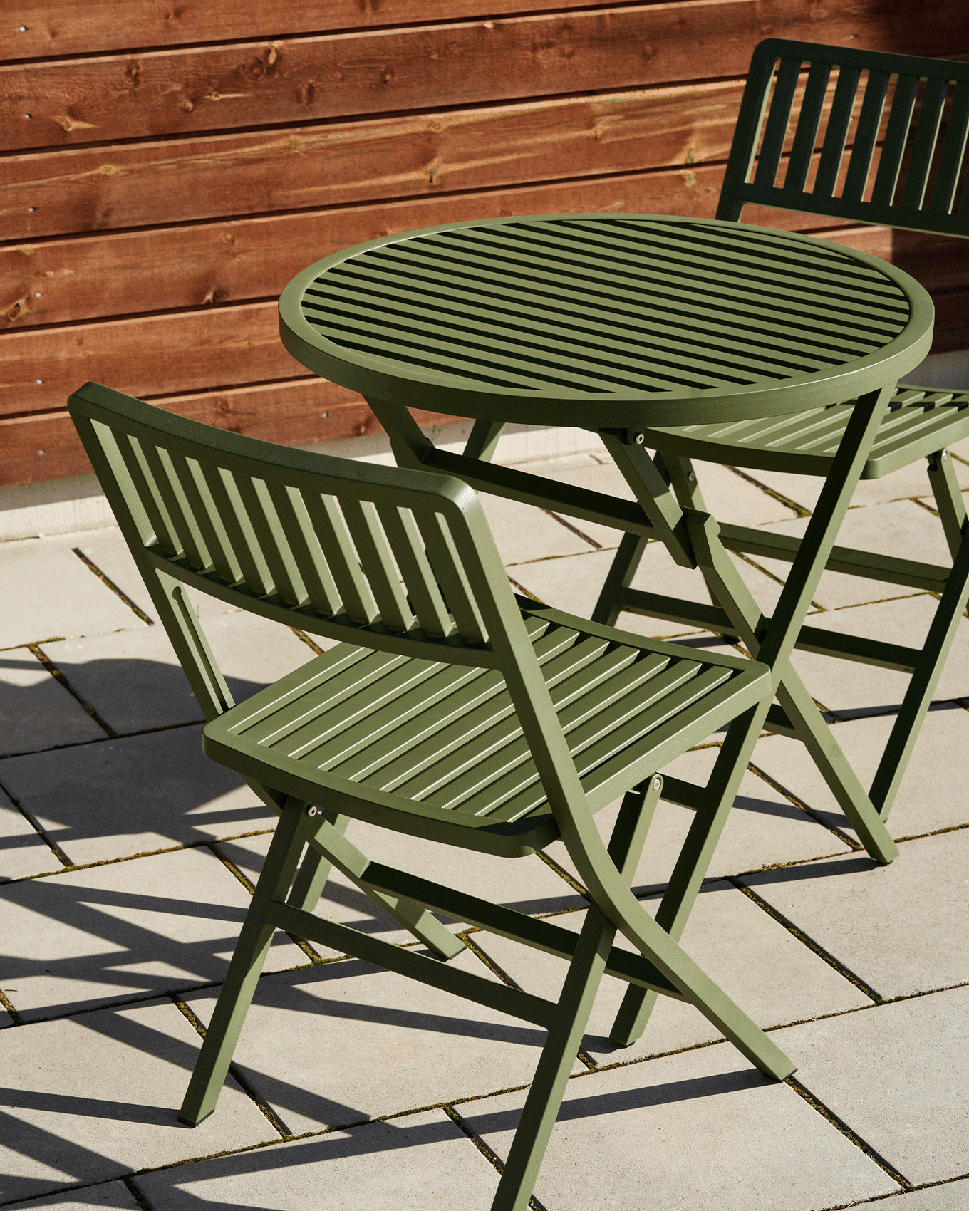 Groene latten tuintafel en twee bijpassende klapstoelen op een terras met een houten schutting.
