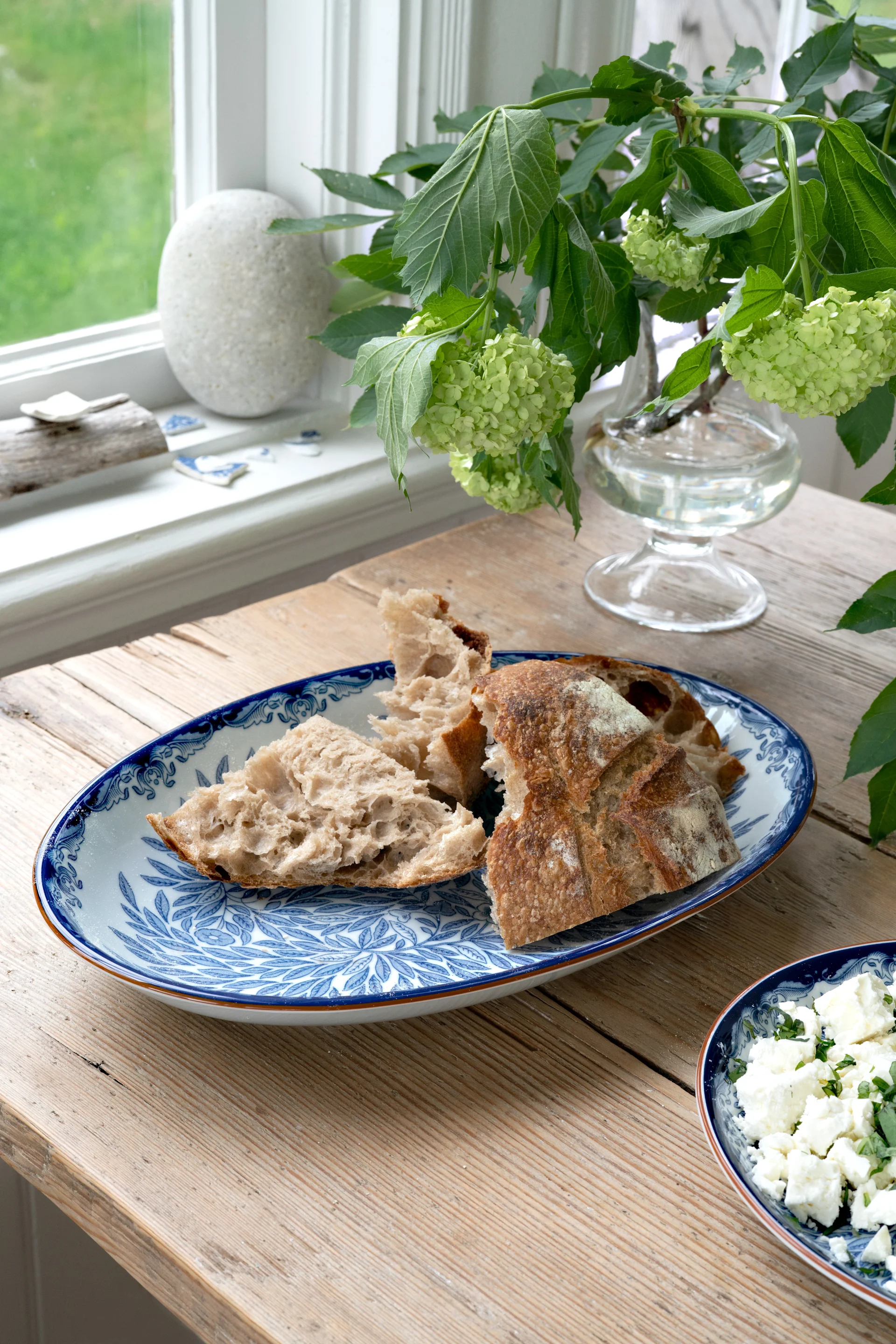 Gescheurde stukken rustiek brood op een blauw-wit gedecoreerd bord, met verkruimelde kaas en groene hortensia's op een houten tafel.