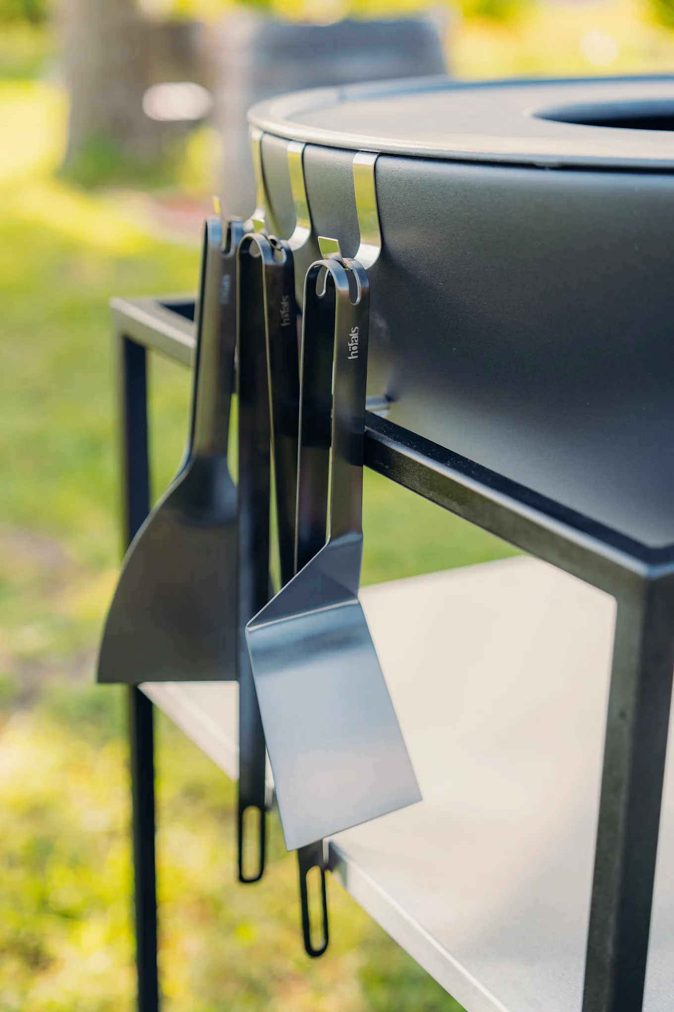 Close-up van een zwarte buitenbarbecue met drie zwarte höfats spatels die aan de zijkant hangen in een tuinomgeving.