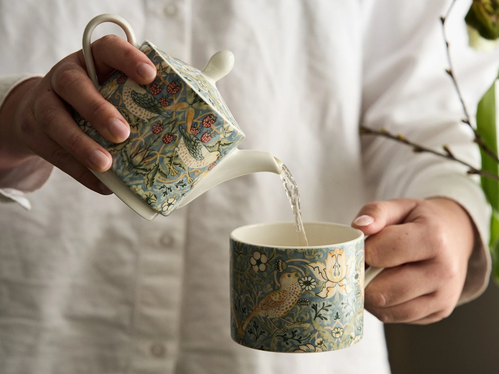 Handen in wit overhemd schenken uit een vierkante theepot met patroon in een bijpassende mok met vogel- en bloemendesign.