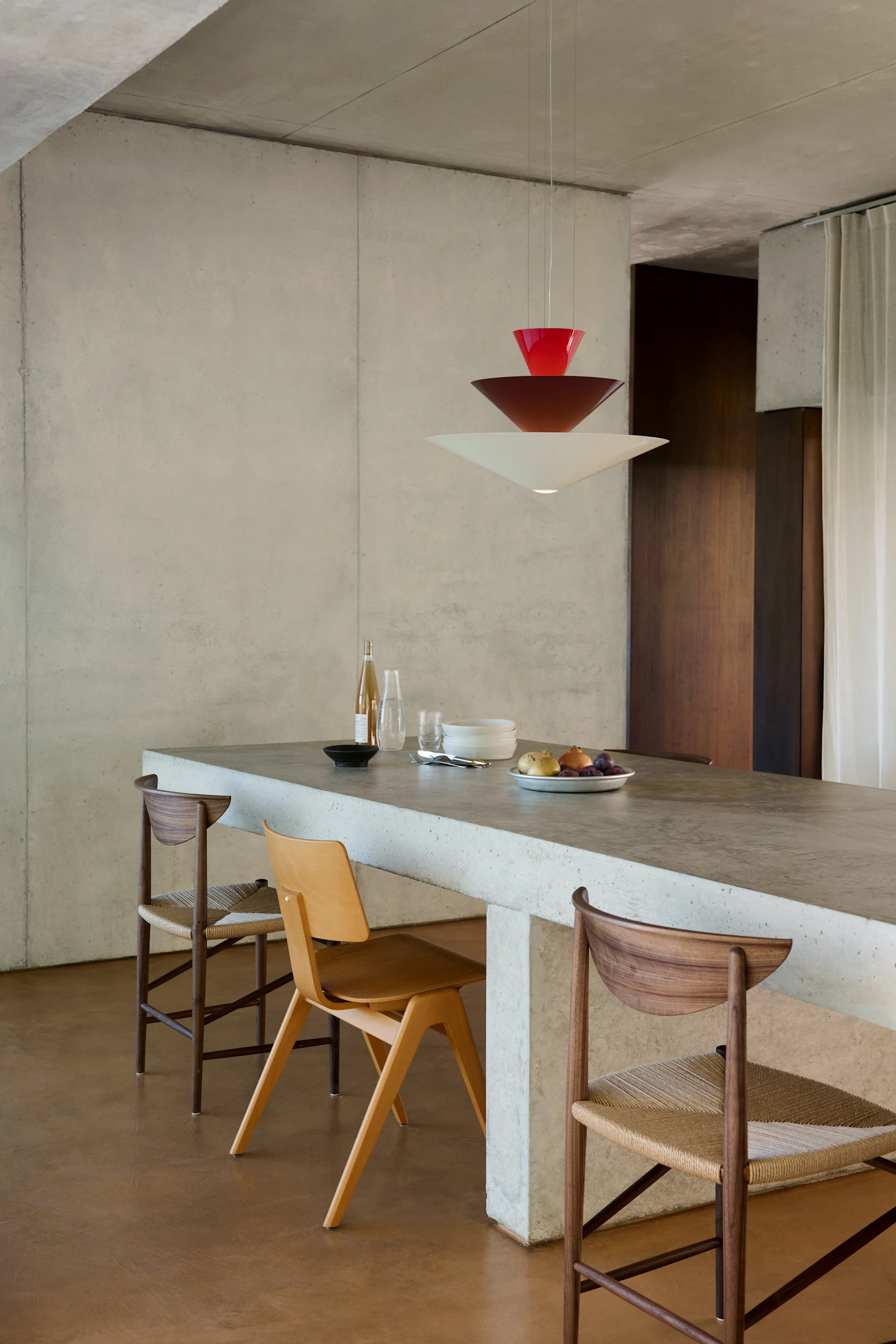 Moderne eethoek met een betonnen eiland, drie houten stoelen en een gelaagde rode en witte hanglamp.