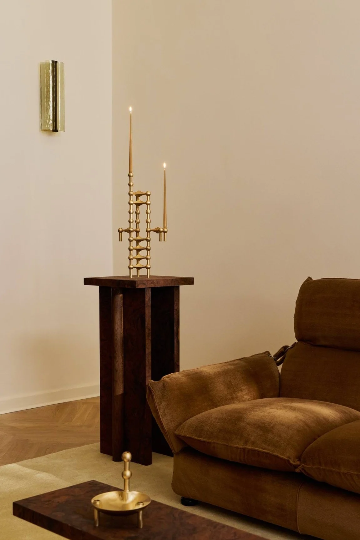 Een sculpturale messing kandelaar met twee brandende kaarsen op een donkere houten sokkeltafel, naast een bruine fluwelen bank.