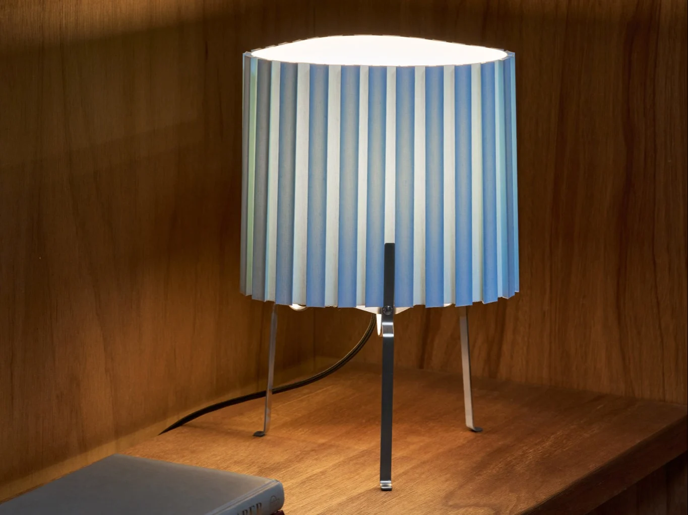Tafellamp met geplooide blauw-wit gestreepte kap op een houten plank, die de omgeving verlicht.