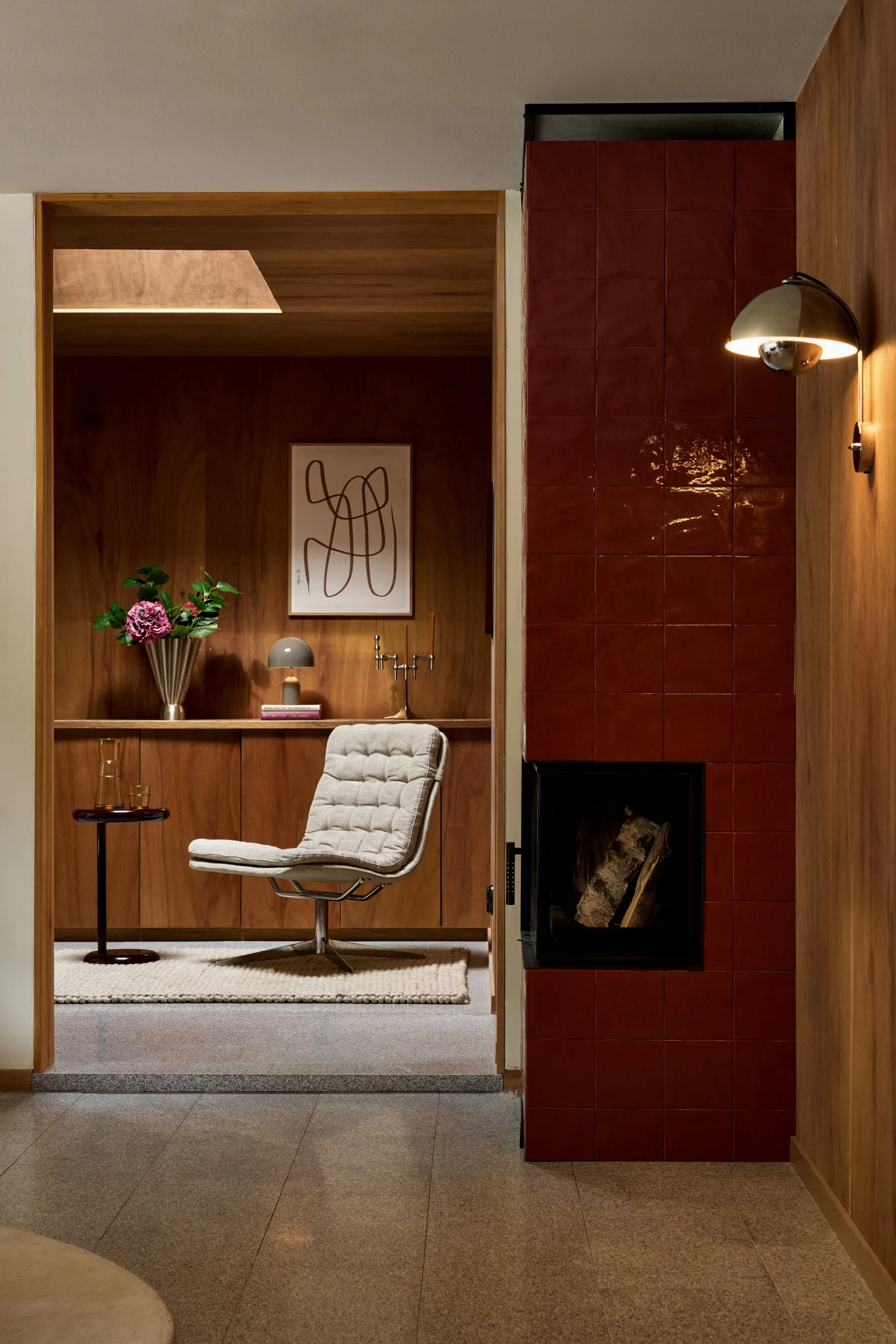 Een gezellig interieur met een gestoffeerde loungefauteuil, houten lambrisering, een rood betegelde open haard en een gouden wandlamp.