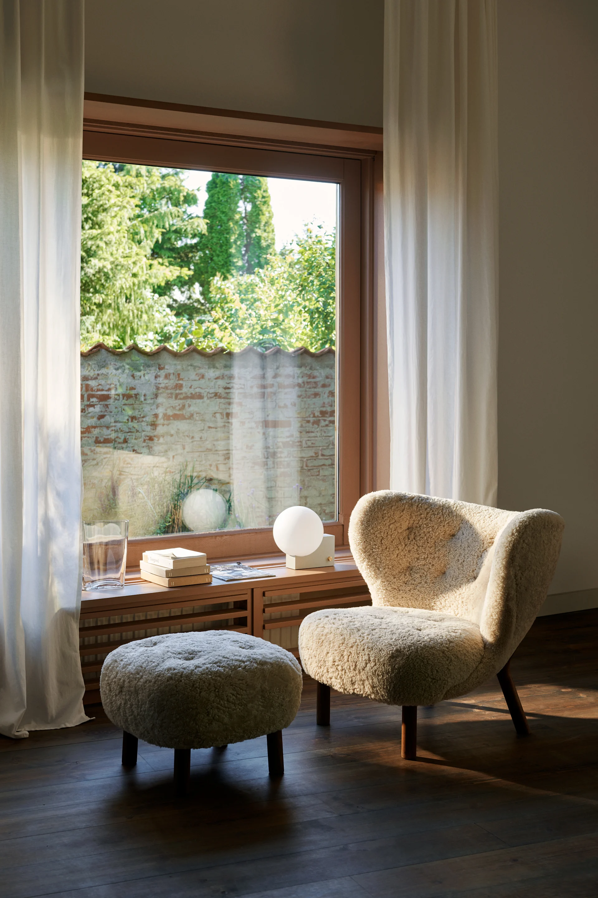 Crèmekleurige bouclé fauteuil en poef bij een zonnig raam. Op de houten vensterbank: een glas, boeken en een sferische lamp.