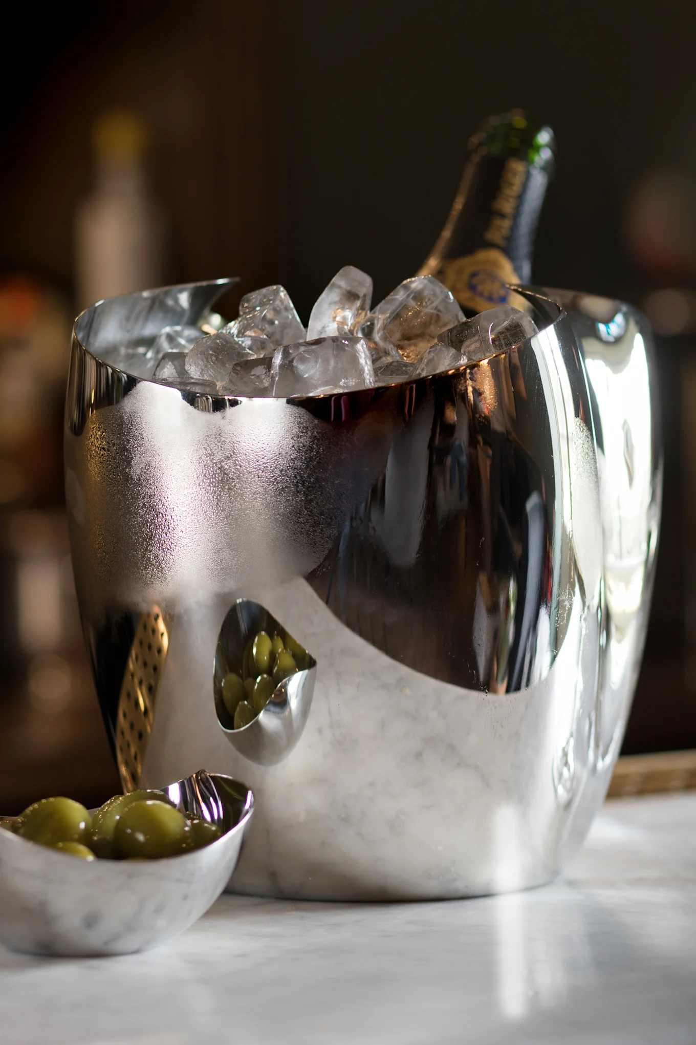 Een reflecterende zilveren ijsemmer met een champagnefles en ijs, naast twee kleine schaaltjes met groene olijven op marmer.