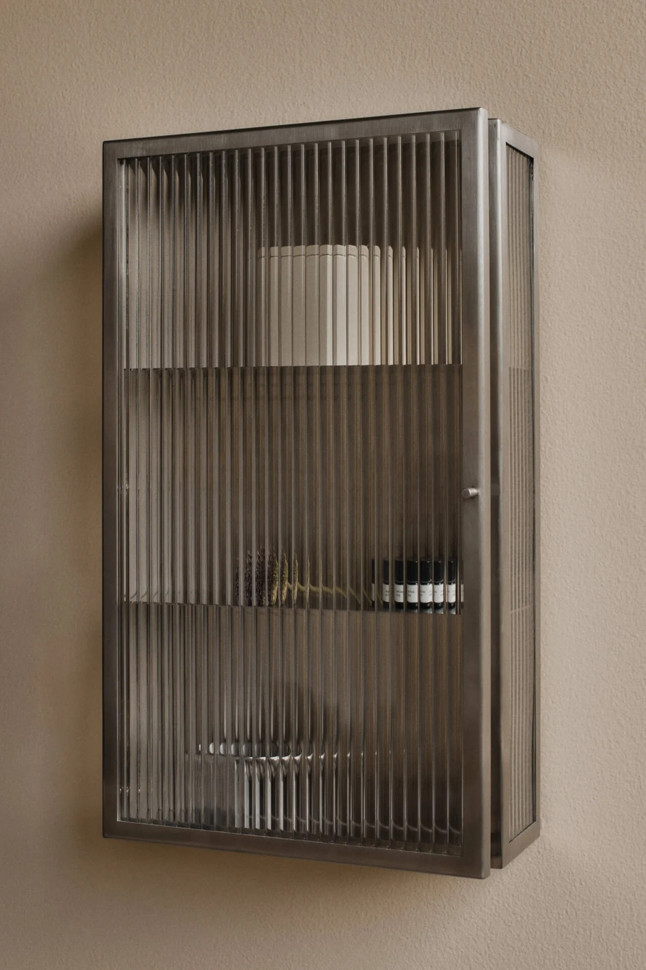 Wandgemonteerde, donkere metalen kast met geribbelde glazen deuren op een beige muur. Bevat planken met een witte cilinder, gedroogde planten en kleine donkere flesjes.