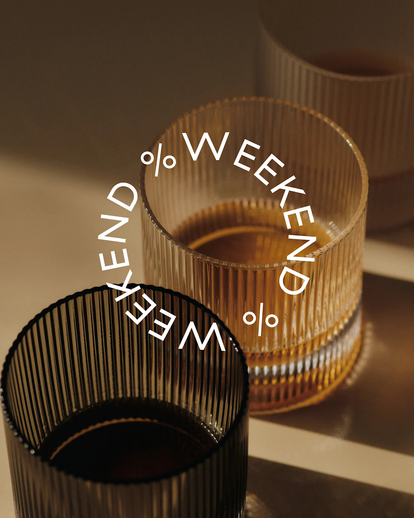 Drie geribbelde glazen, twee gevuld met donkere vloeistof. Een donker glas vooraan, een amberkleurig glas erachter. Tekst: Weekend %.