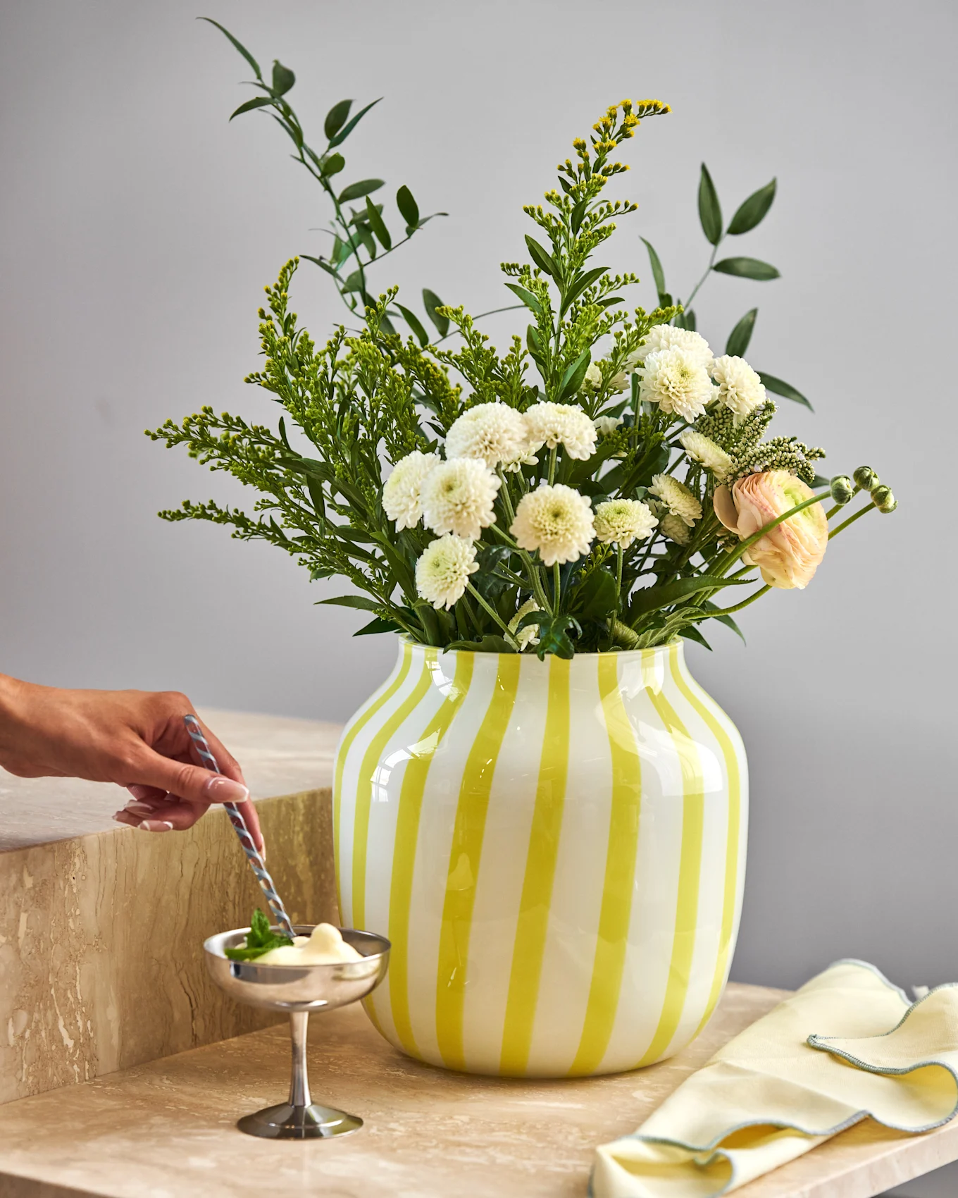 Hand roert dessert in zilveren kom op marmer. Geel gestreepte vaas met witte chrysanten, gele bloemen en roze ranonkel.