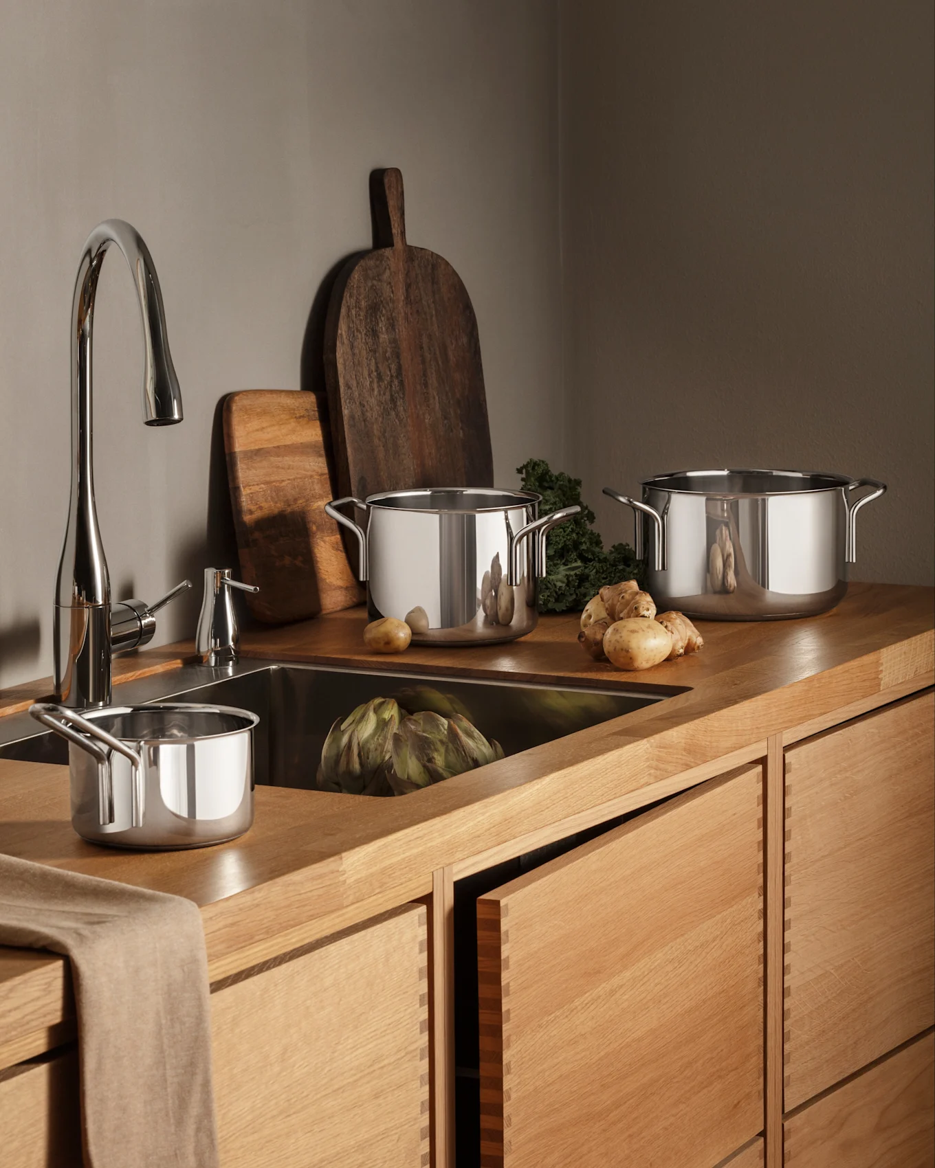 Een moderne keuken met houten werkbladen en kasten, een chromen kraan en roestvrijstalen pannen, verse producten waaronder artisjok en aardappelen.