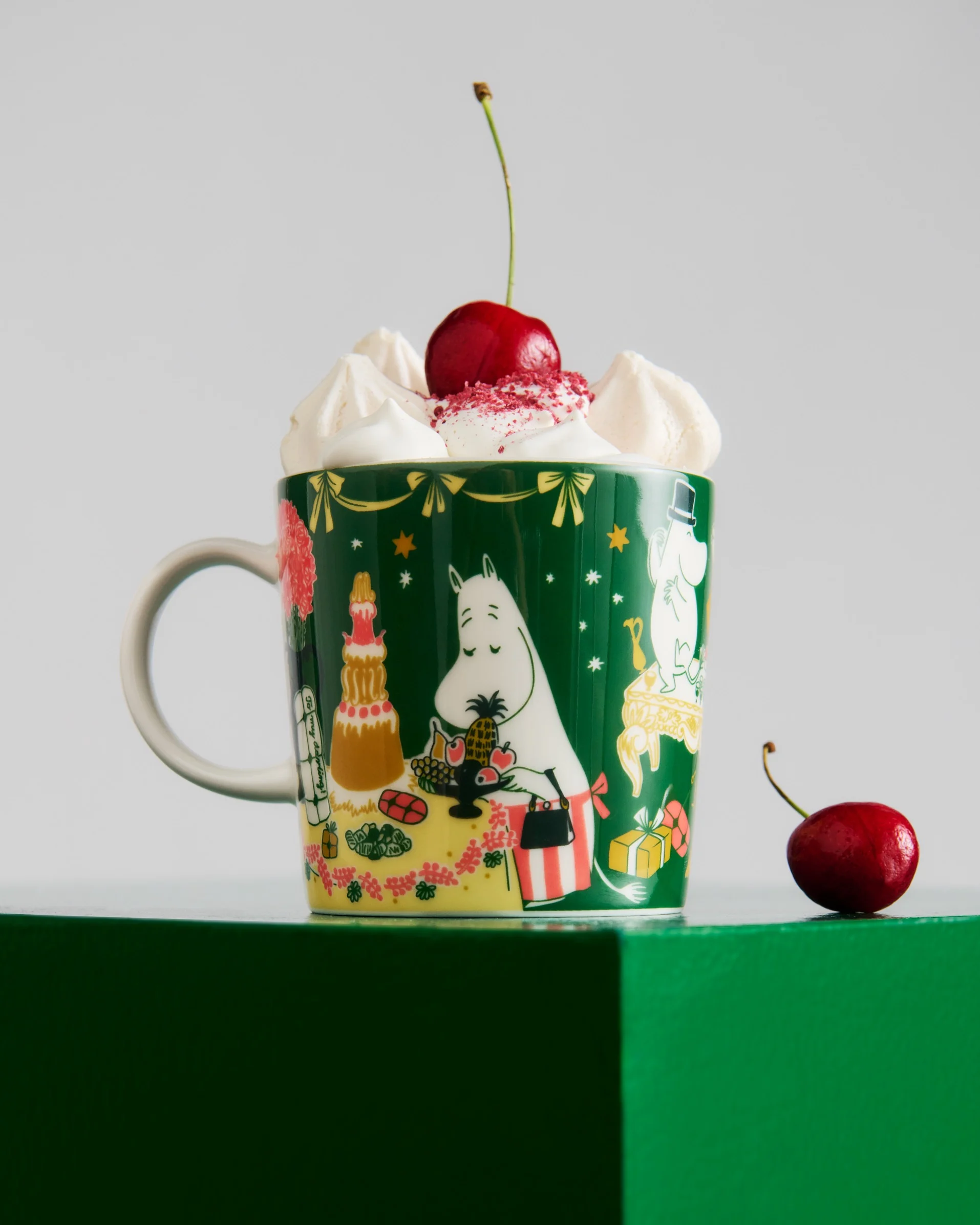 Donkergroene mok met Moomin-personages, gevuld met slagroom, rode hagelslag en een kers. Een andere kers ligt op het groene oppervlak.