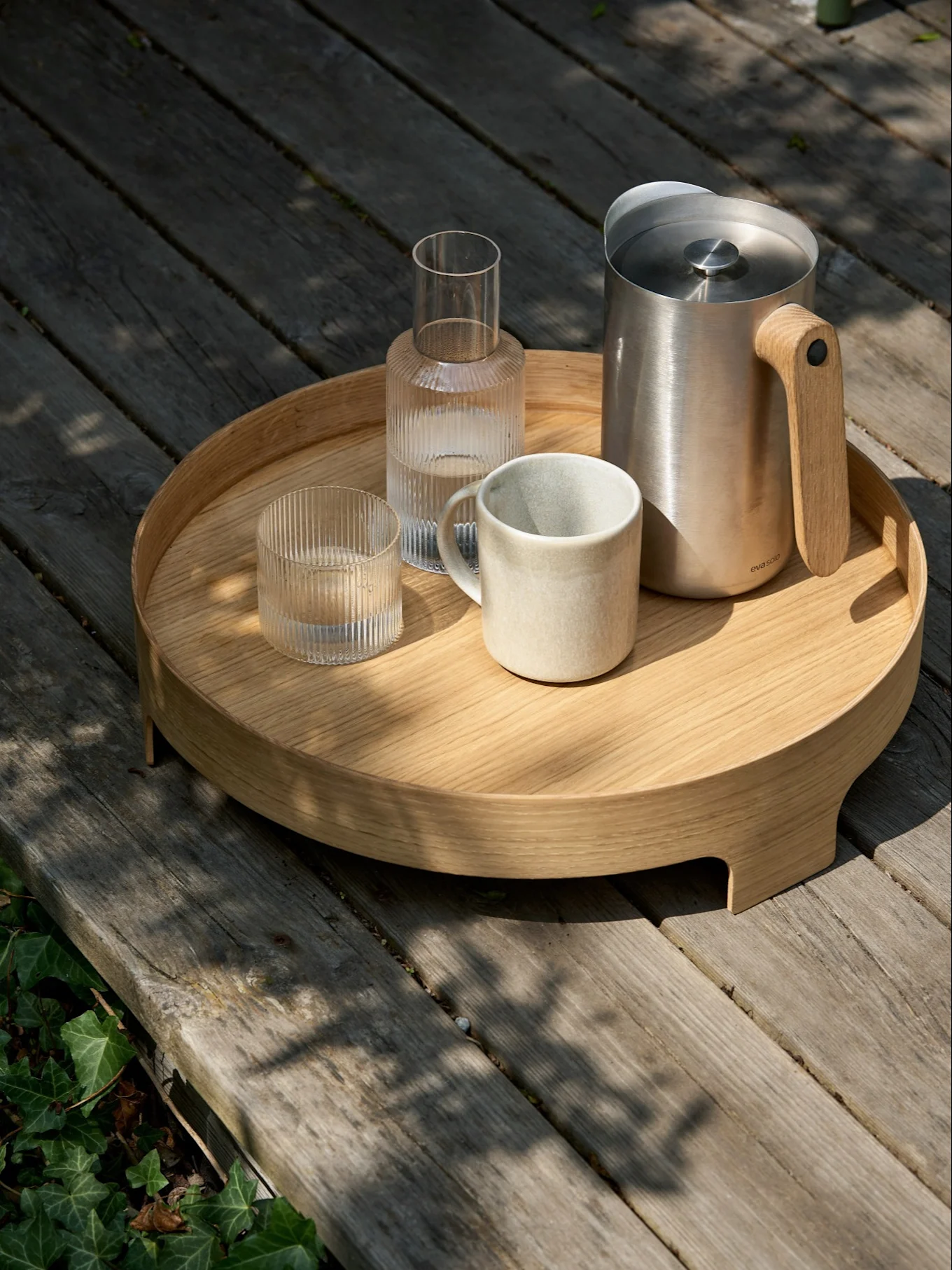 Rond houten dienblad op een terras met roestvrijstalen kan, keramische mok en twee geribbelde glazen karaffen.